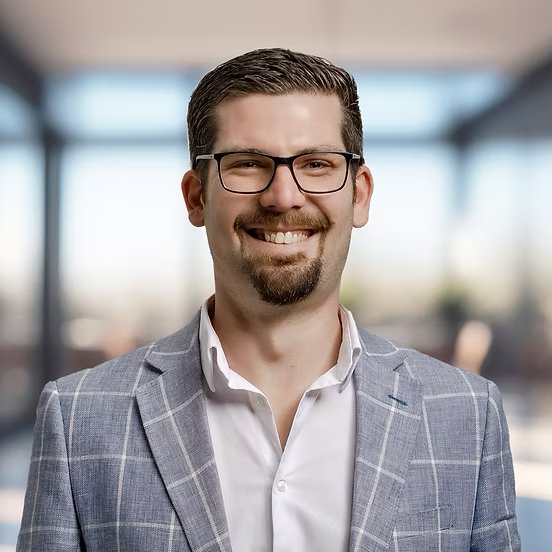 A smiling man wearing glasses, a white shirt, and a gray checkered blazer standing indoors with a blurred background.