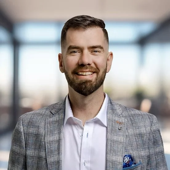 A smiling man with a beard and short hair wearing a light gray plaid blazer and white dress shirt, standing in a modern office with large windows in the background.