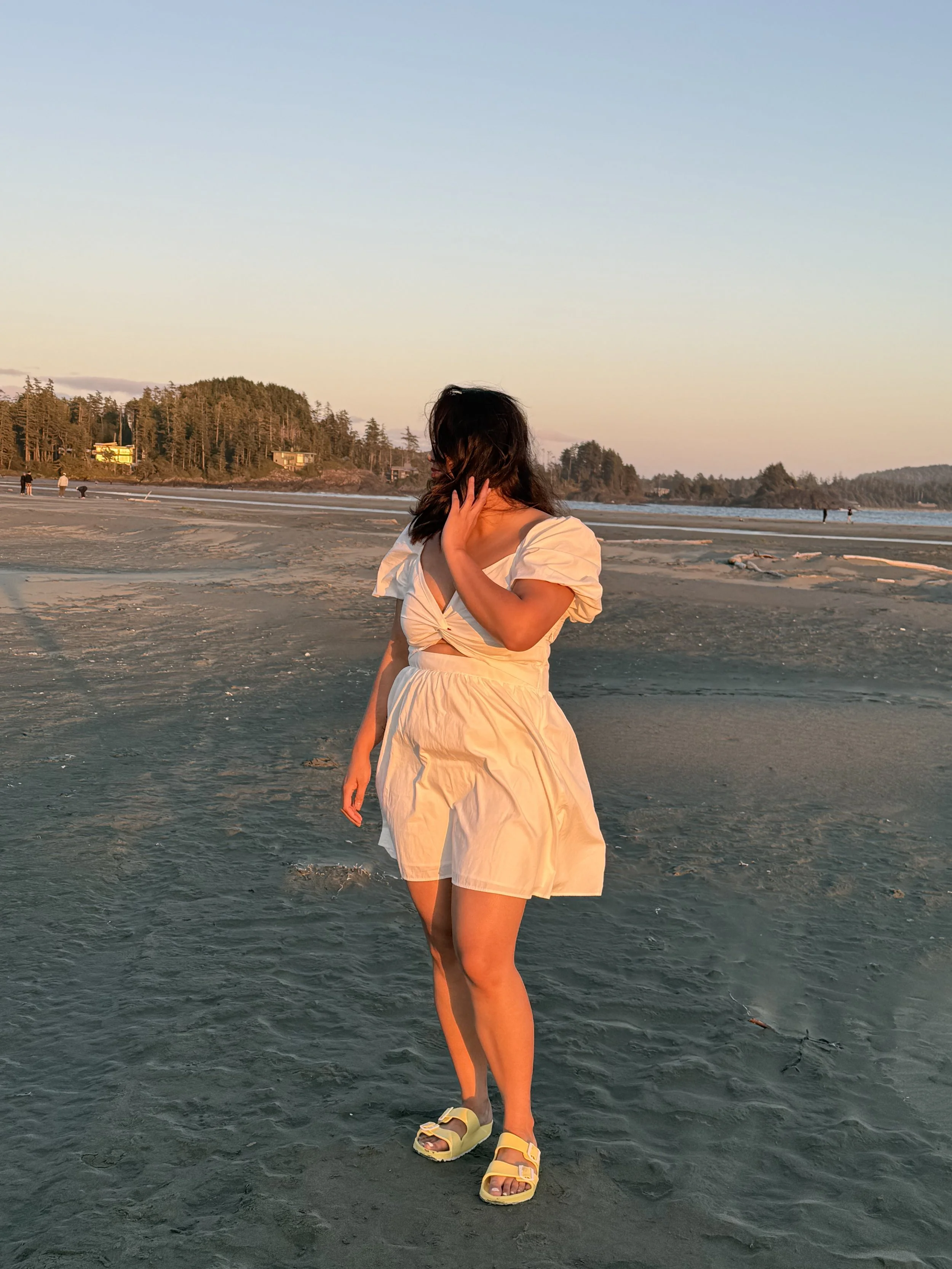 A woman in a white dress and sandals walking on a sandy beach during sunset, with trees and small buildings in the background.