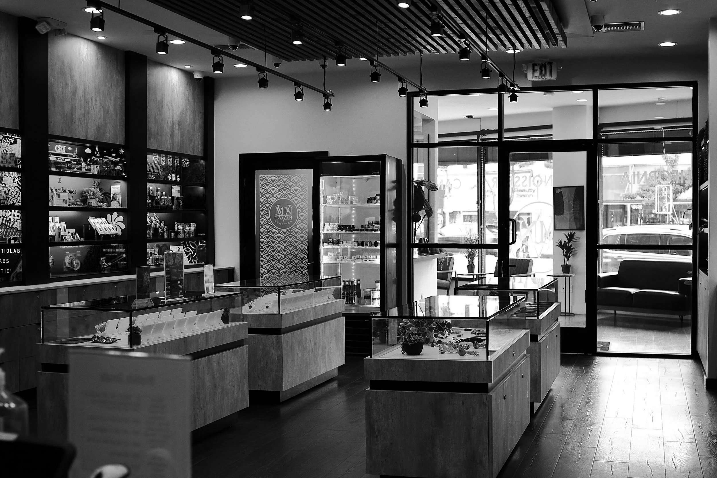 Black and white photo of a retail store interior with display cases, shelves with products, a vending machine, seating area near large glass doors, and track lighting on the ceiling.
