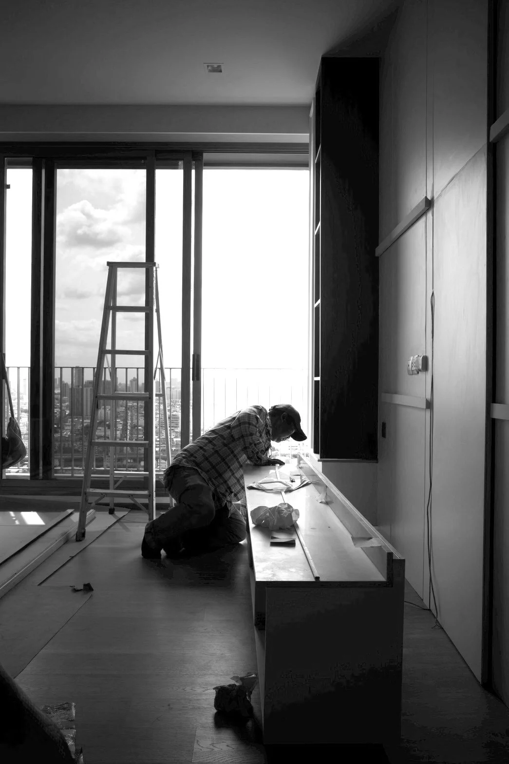 A construction worker kneeling on the floor, working on a piece of furniture in a high-rise apartment during the daytime, with a city skyline visible through large sliding glass doors and an unfinished ladder nearby.