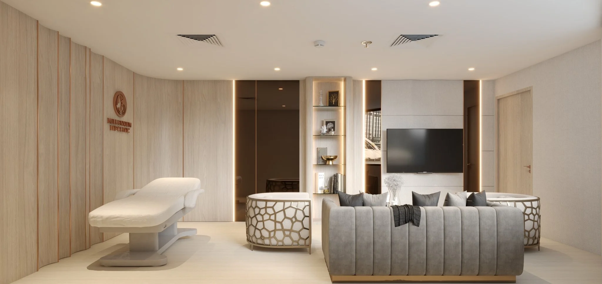 Interior of a modern medical examination room featuring a reclining examination chair, a gray velvet sofa with black and gray pillows, a large flat-screen TV, and built-in shelves with decorative items. The room has a light wood-paneled wall with a healthcare logo and gentle LED lighting.