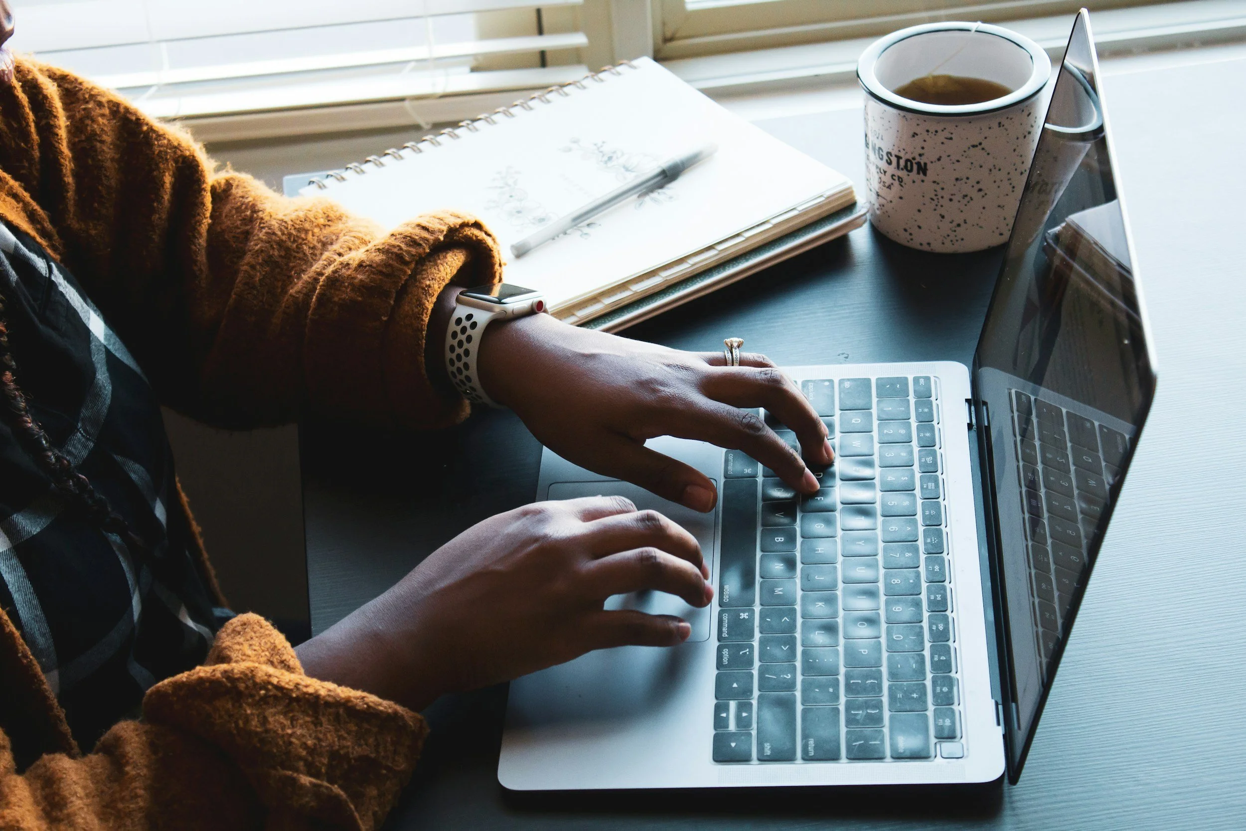 A person working on a laptop at a desk, with a cup of coffee, a sketchbook, and a pen nearby.