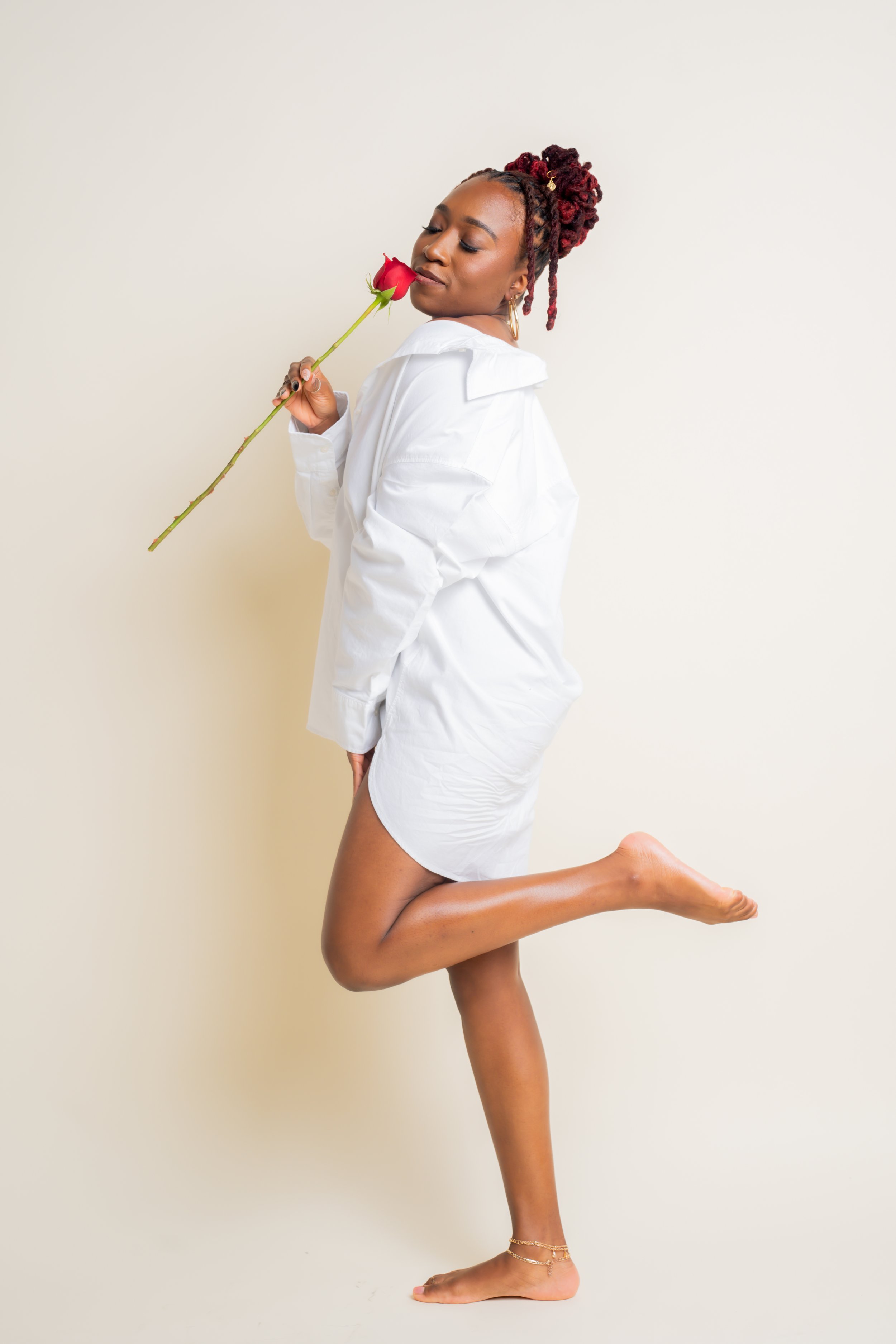 A woman with red dreadlocks holding a red rose to her nose, wearing a white oversized shirt and barefoot, standing against a plain light-colored background.