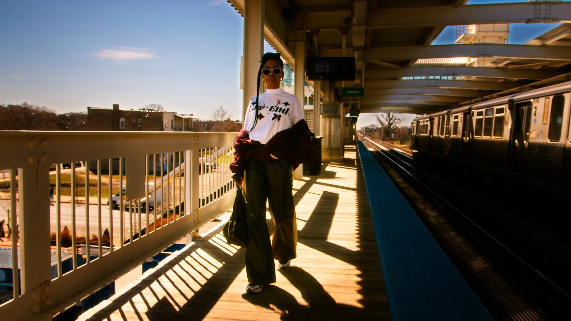 A woman with sunglasses walking on a train station platform during daytime, with a train in the background and the sun casting shadows.