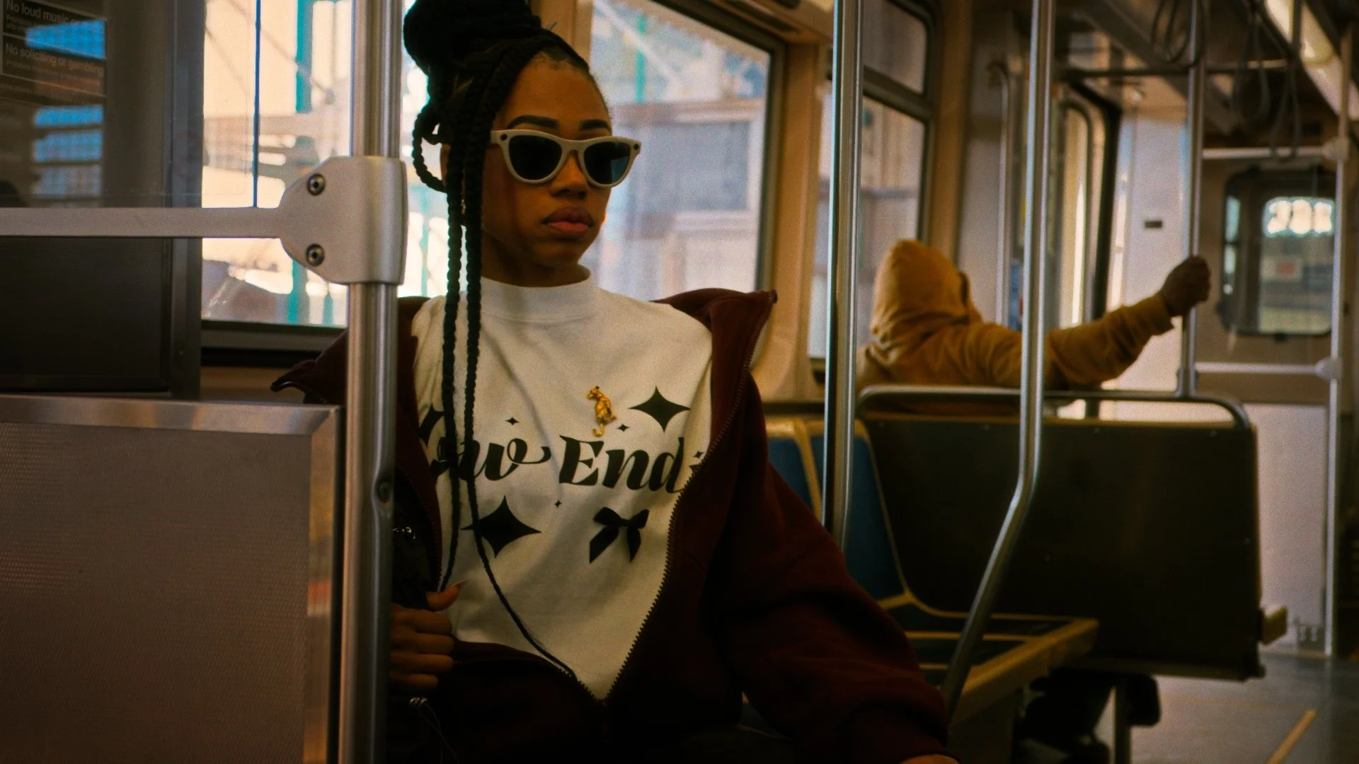 A young woman with braided hair, wearing white sunglasses, a white t-shirt with black graphics and text, and a brown jacket, sitting on a bus. She has a small gold pin on her shirt and earphones in. Another passenger in a yellow hoodie is seated behind her, leaning back with their arm extended with their fist in the air.