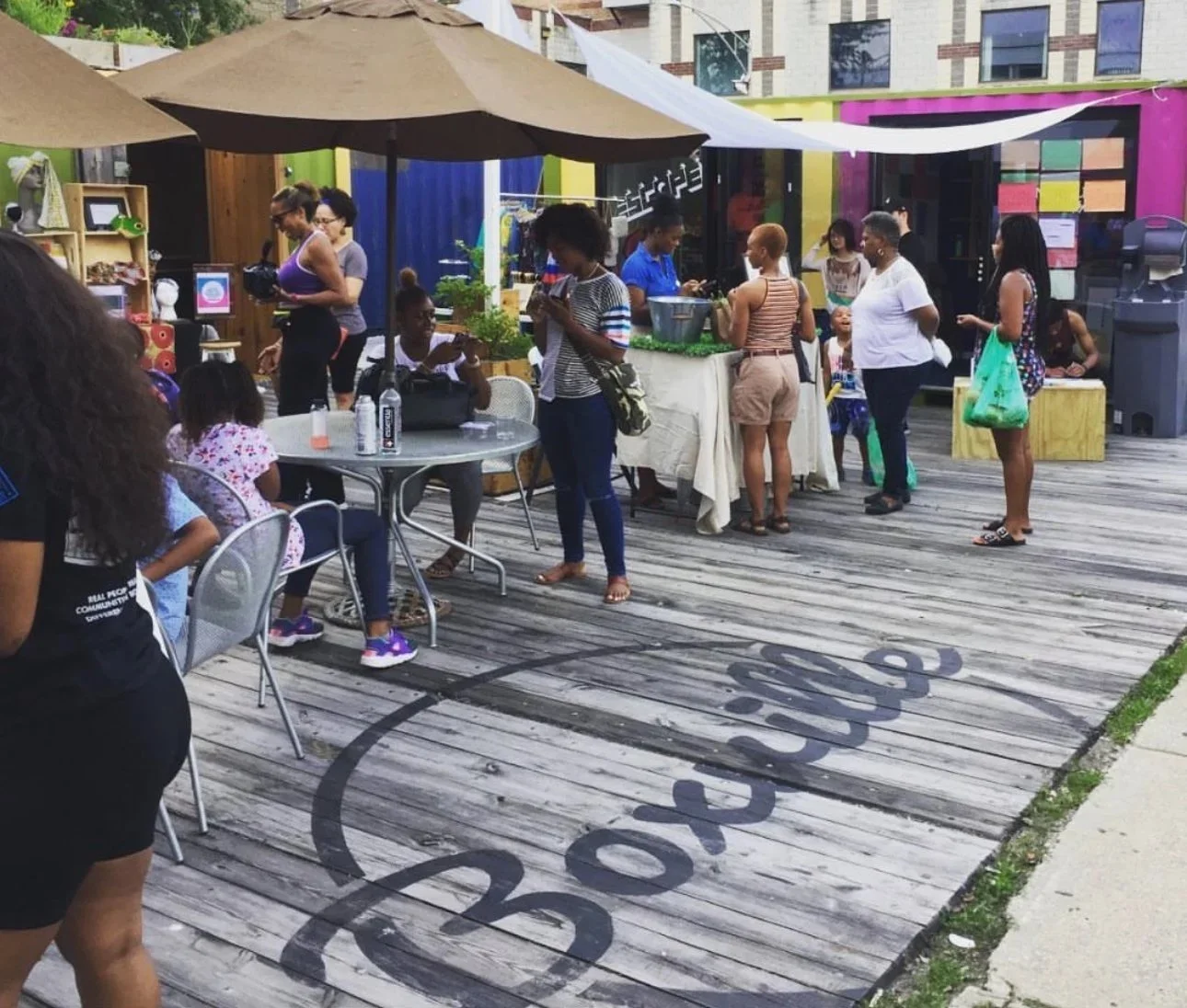 People at an outdoor market or community event on a wooden deck with umbrellas and colorful building in the background.