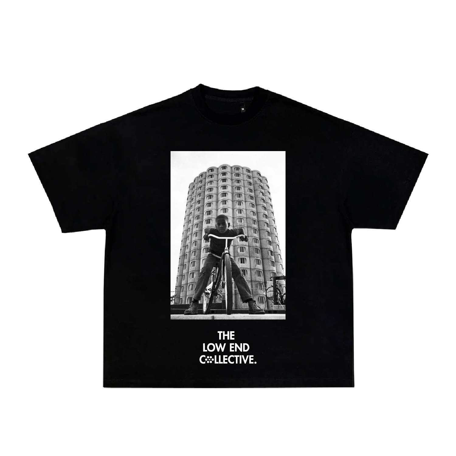 Black T-shirt featuring a black-and-white photo of a boy on a bicycle in front of a tall, rounded apartment building. The shirt has the text "The Low End Collective" below the photo.