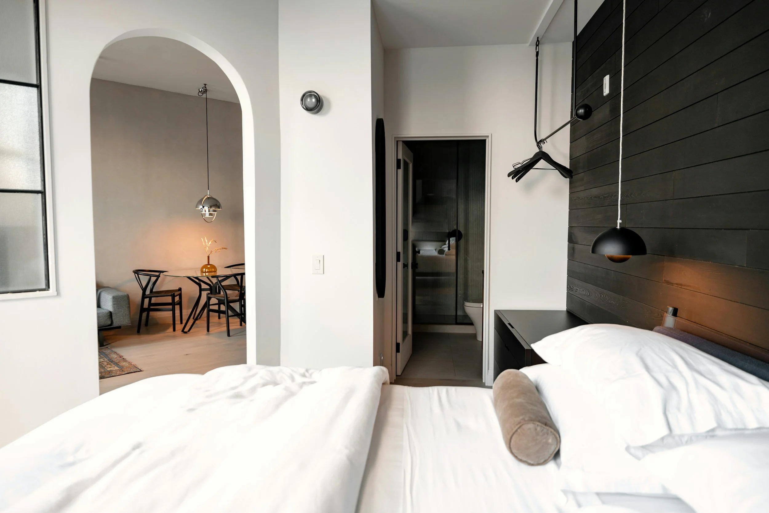 A bedroom with white bedding and pillows, a dark wooden headboard with a hanging black lamp, a small black side table, an avid hanging above a dark accent wall, and a small open bathroom with a toilet visible. An arched doorway leads to a dining area with a round table, four black chairs, a pendant light, and a vase with dried flowers.