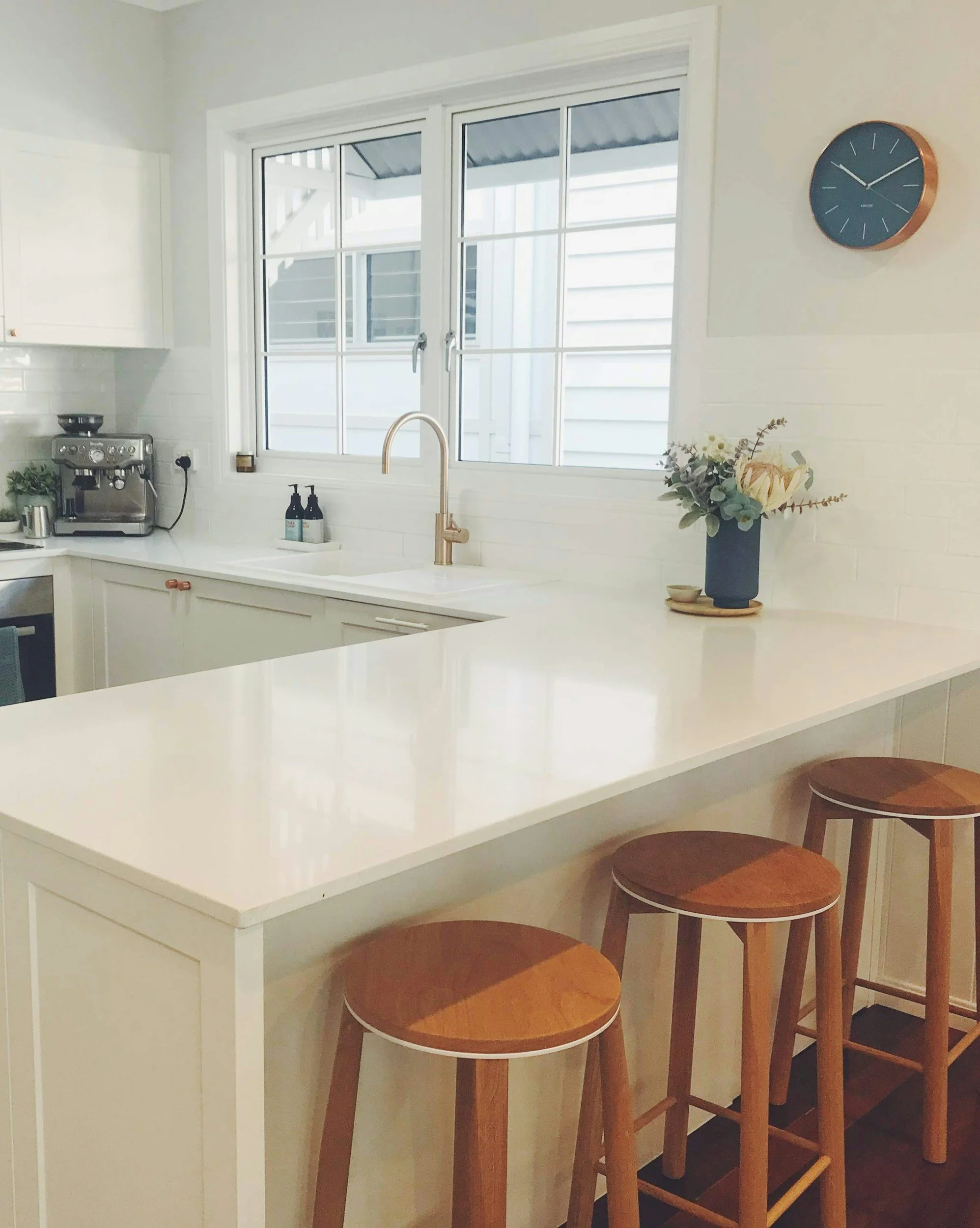 Clean, white kitchen with a large window, a white countertop, a sink with a gold faucet, three wooden barstools, a clock on the wall, and a vase of flowers on the counter.