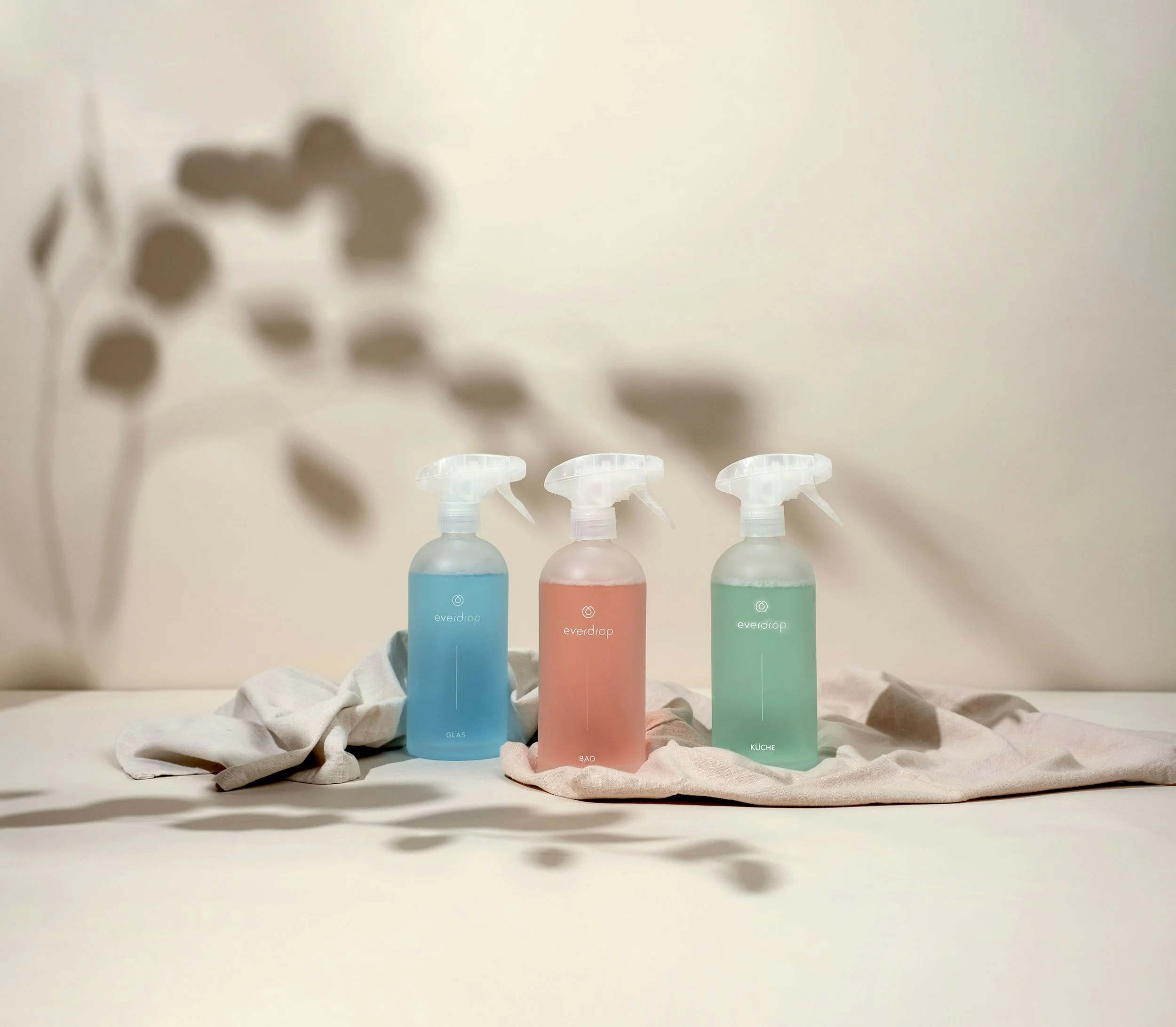 Three spray bottles with pastel liquids labeled 'GLAS,' 'BAD,' and 'KUCHE,' placed on a light cloth with shadows of a plant on the wall in the background.