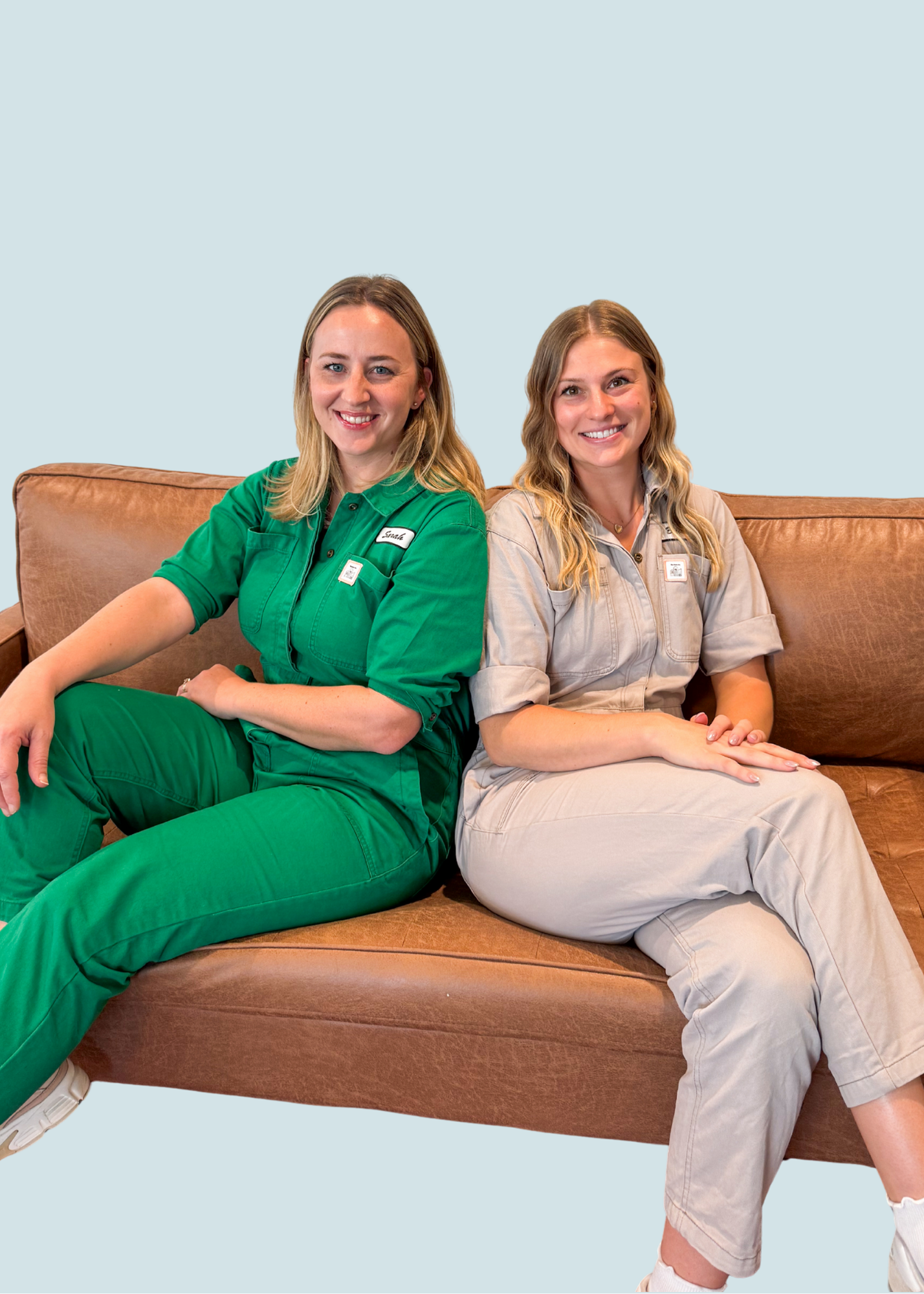 Two women sitting on a brown couch, one in green work uniform and the other in beige work uniform, smiling at the camera, against a light blue background.