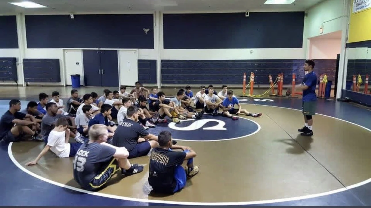 A group of young boys and teenagers sitting on the floor of a wrestling gym, attentively listening to a coach who is standing in front of them. The gym has black and gold markings on the floor and various training equipment along the walls.