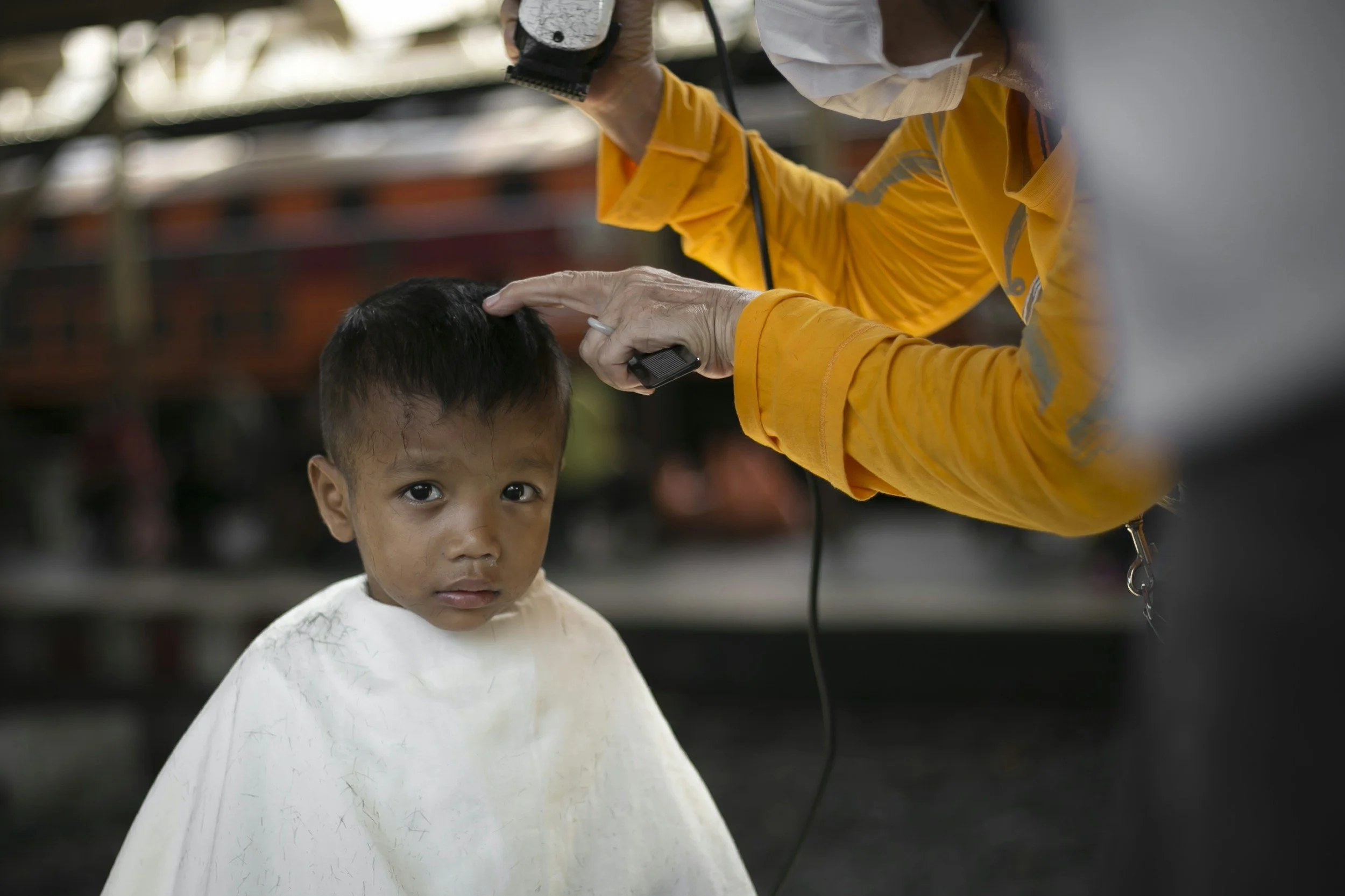 A young boy with a white haircut and dirt on his face sits in a barber's chair while a barber trims his hair using electric clippers.