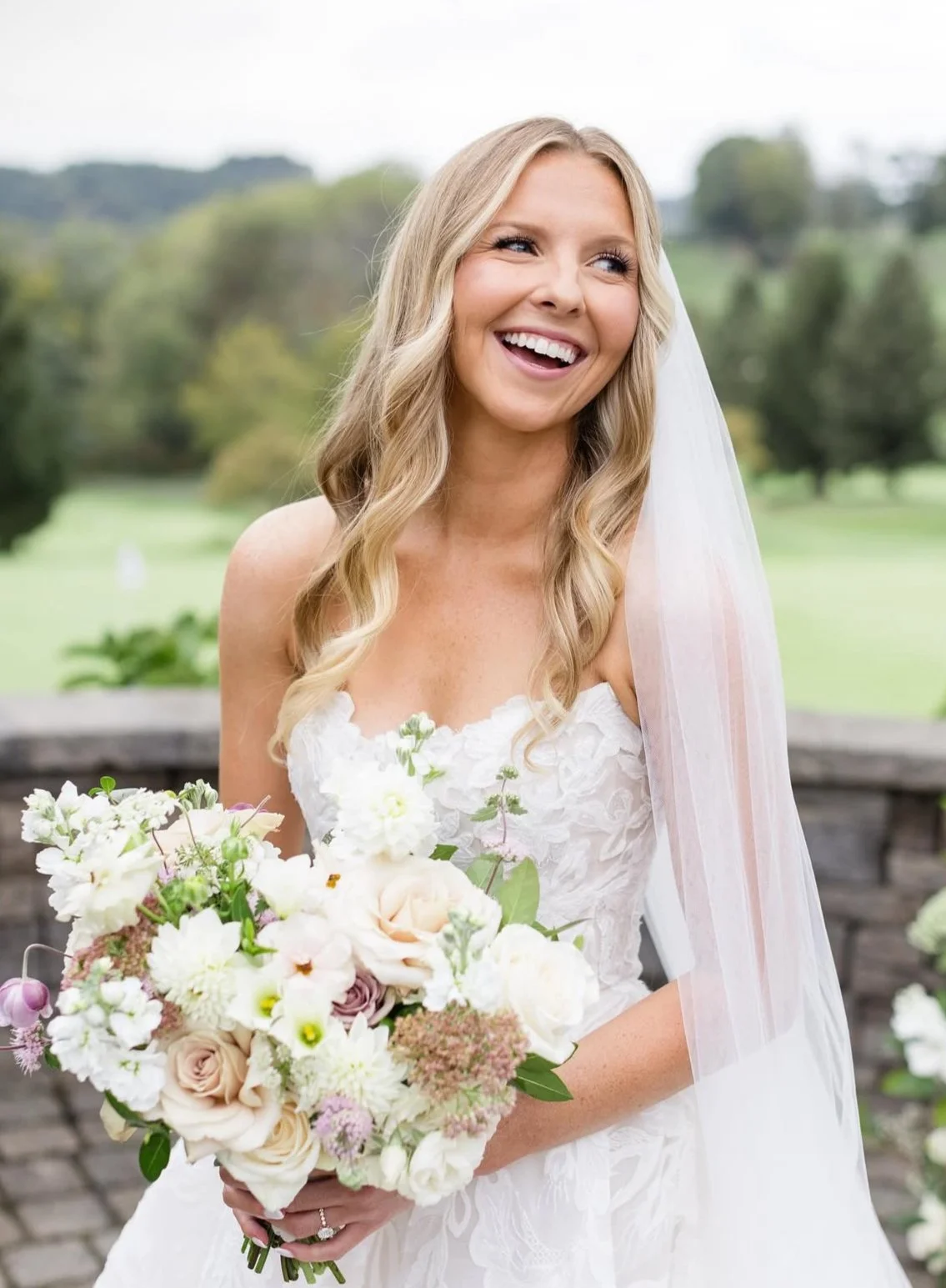 Blonde bride wearing a long white veil and holding a bouquet of white, beige, and purple flowers with green details and blurred trees and grass in the background