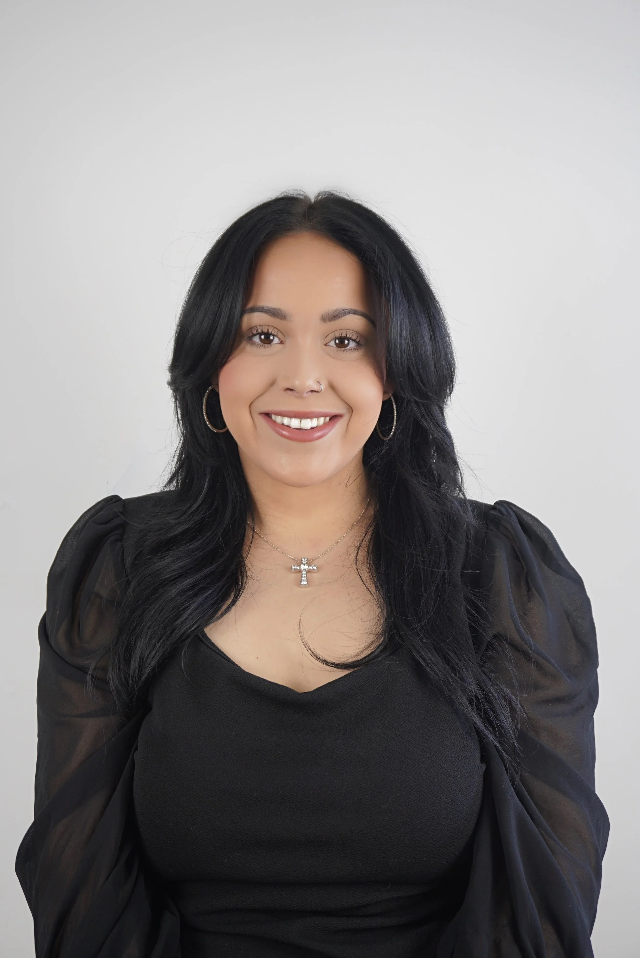 A woman with long, wavy black hair, wearing a black top with puffed sleeves, silver hoop earrings, a necklace with a cross pendant, and a nose piercing, smiling against a plain white background.