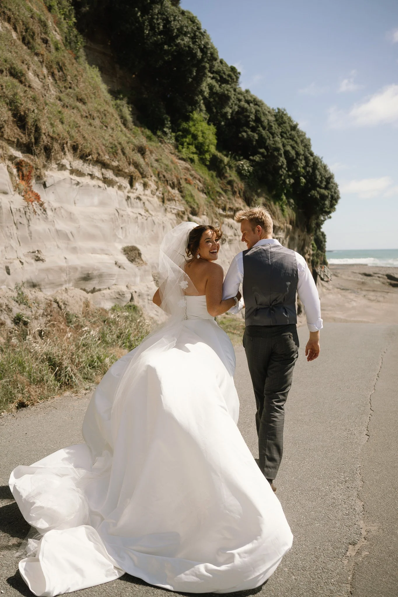 A bride and groom walking hand in hand along a coastal road with cliffs and the ocean in the background, smiling and looking at each other.