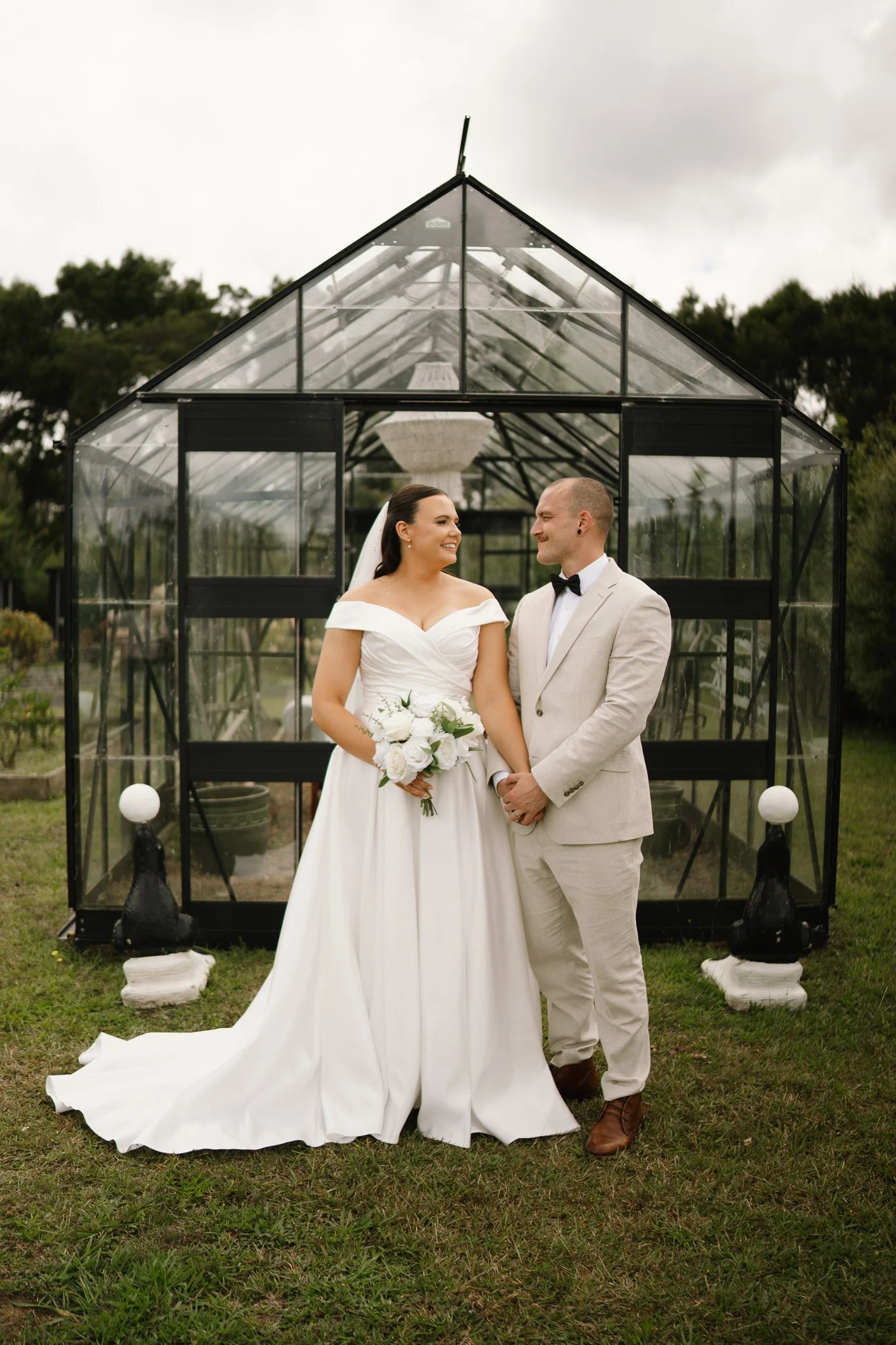 A bride in a white wedding dress holding a bouquet, and a groom in a light-colored suit with a black bow tie, standing outdoors in front of a glass greenhouse, holding hands and smiling at each other.