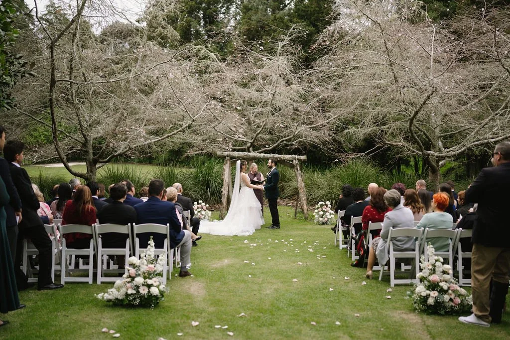 A wedding ceremony outdoors with a bride and groom exchanging vows under a rustic wooden arch. Guests seated on white chairs watch the scene, surrounded by blooming trees and floral arrangements.