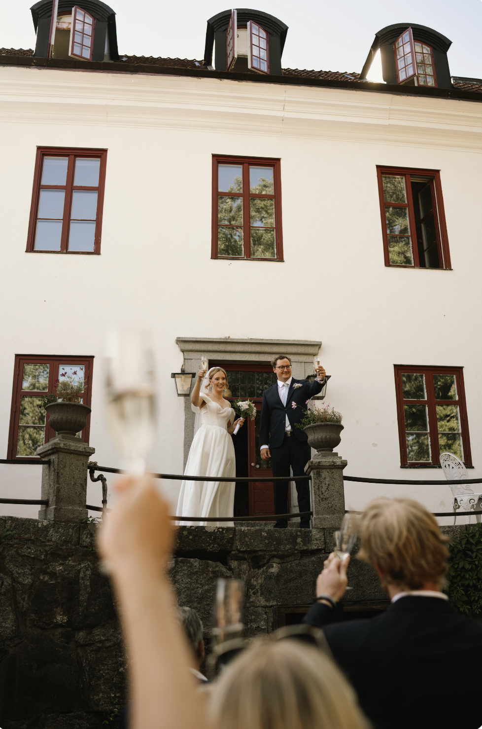 A newlywed couple standing on the front steps of a white house with red window frames, raising glasses in a toast at their wedding celebration, with guests in the foreground raising glasses.