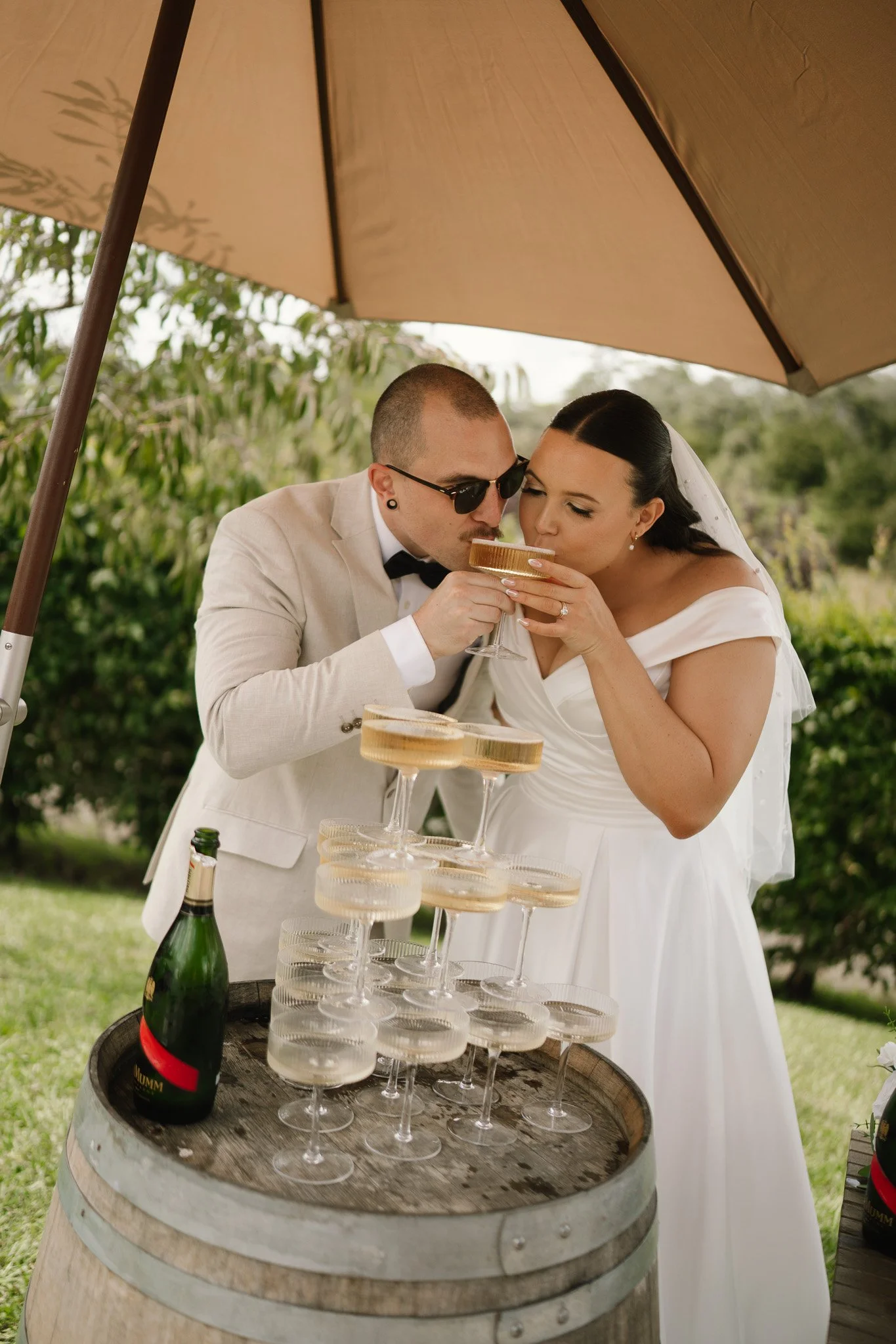 A bride and groom in wedding attire underneath a large beige umbrella, sharing a toast in champagne glasses decorated with gold rims, standing beside a wooden barrel with a bottle of champagne and stacked glasses.