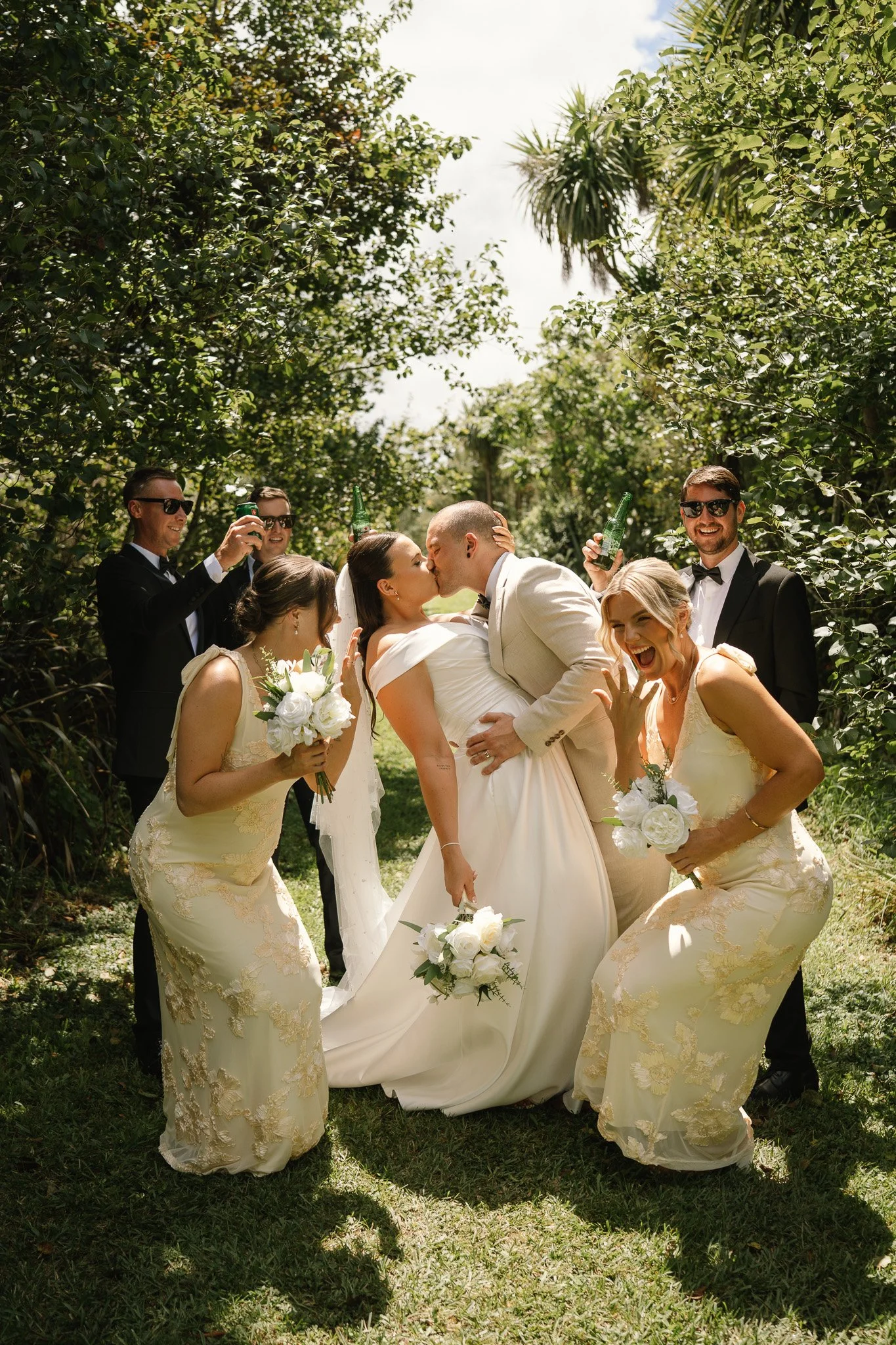 A wedding celebration outdoors with the bride and groom kissing, surrounded by bridesmaids and groomsmen, some holding drinks and flowers, in a lush green setting.