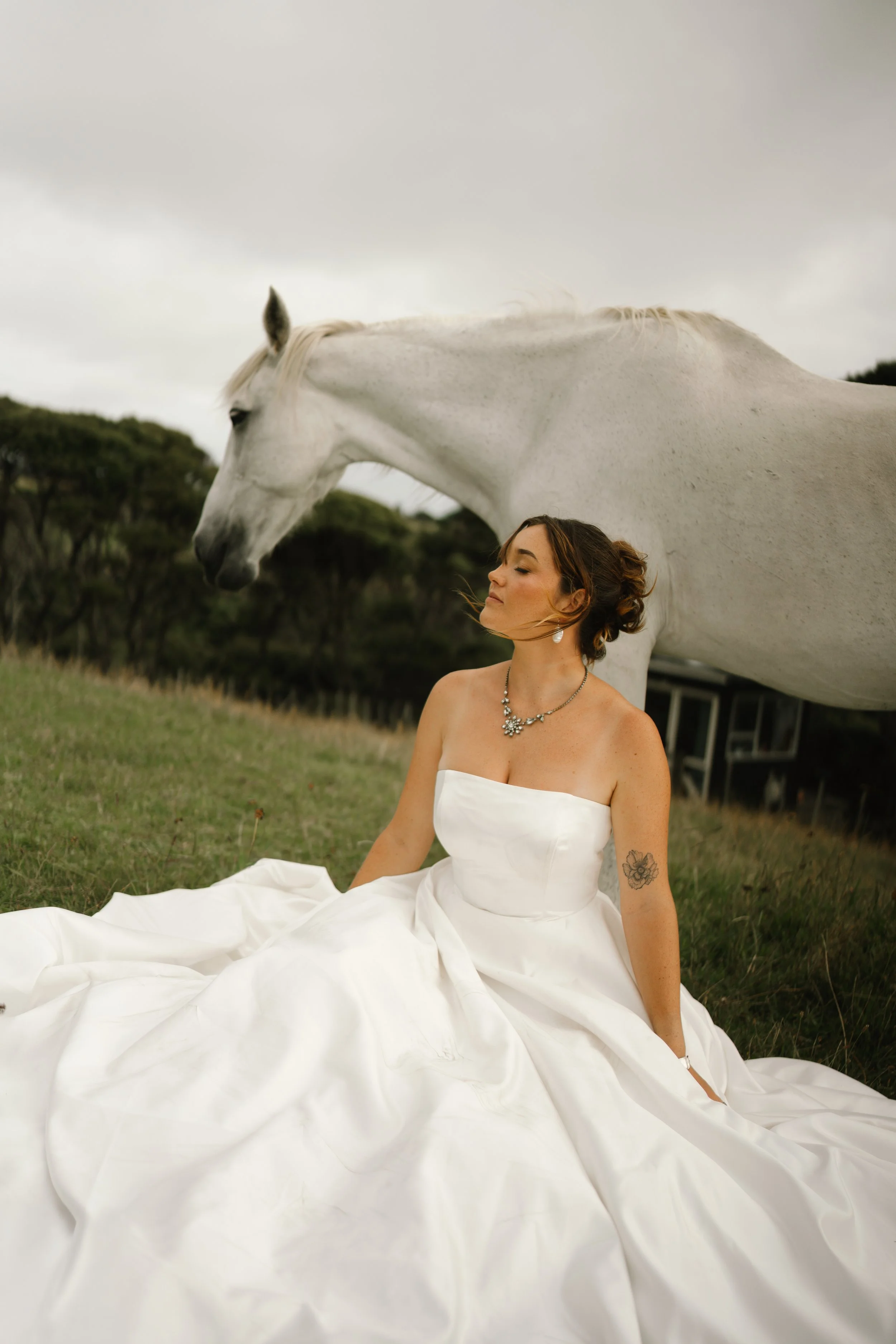 A woman in a white wedding dress sitting on grass, with a white horse standing behind her on a cloudy day, surrounded by trees.