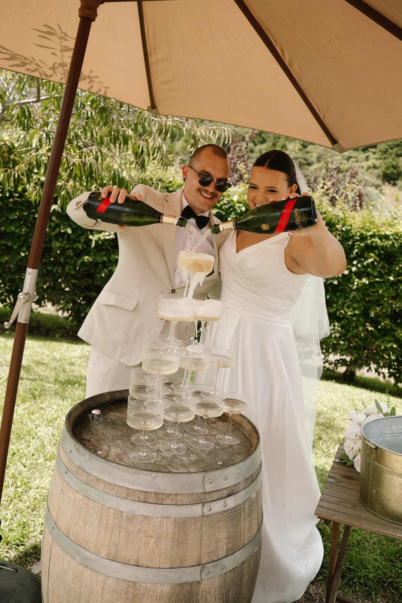 Bride and groom pouring champagne into a pyramid of glasses during their outdoor wedding celebration.