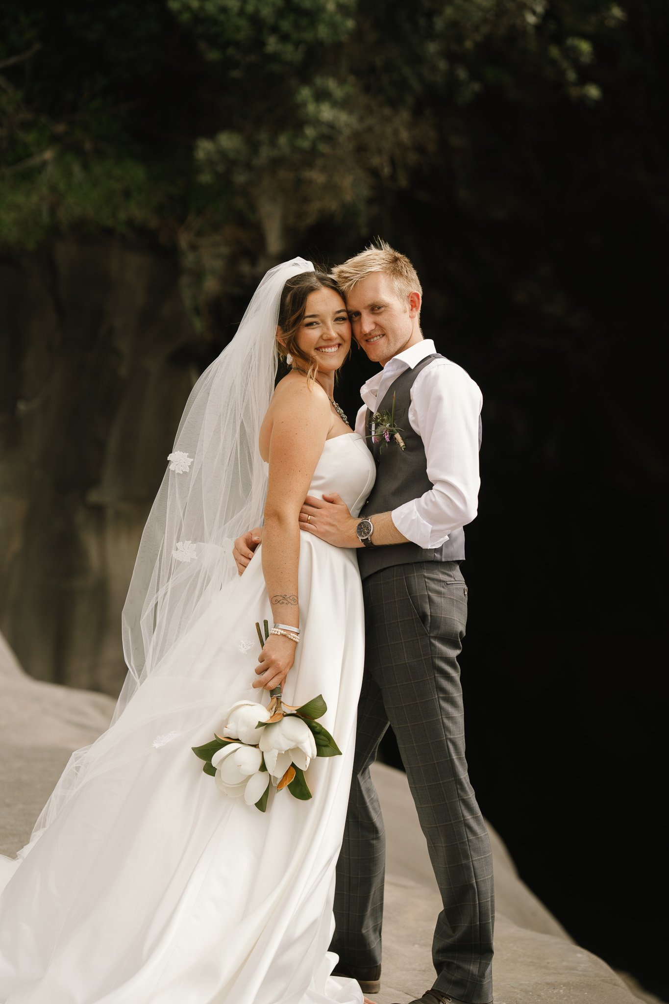A bride and groom standing close together on a beach, smiling, with lush trees and a dark waterfall in the background.