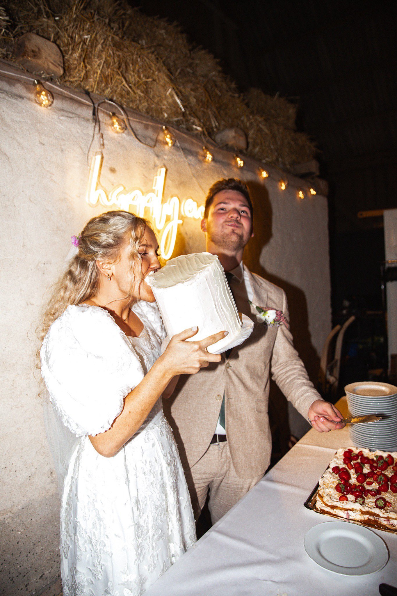 A woman in a white wedding dress is biting into a layered wedding cake with white frosting. A man in a beige suit stands beside her, holding a cake server, at a wedding reception with a white tablecloth, a fruit-topped dessert, and a vertical stack of plates. Pink and white flowers are on his lapel, and there is a neon sign and string lights in the background.