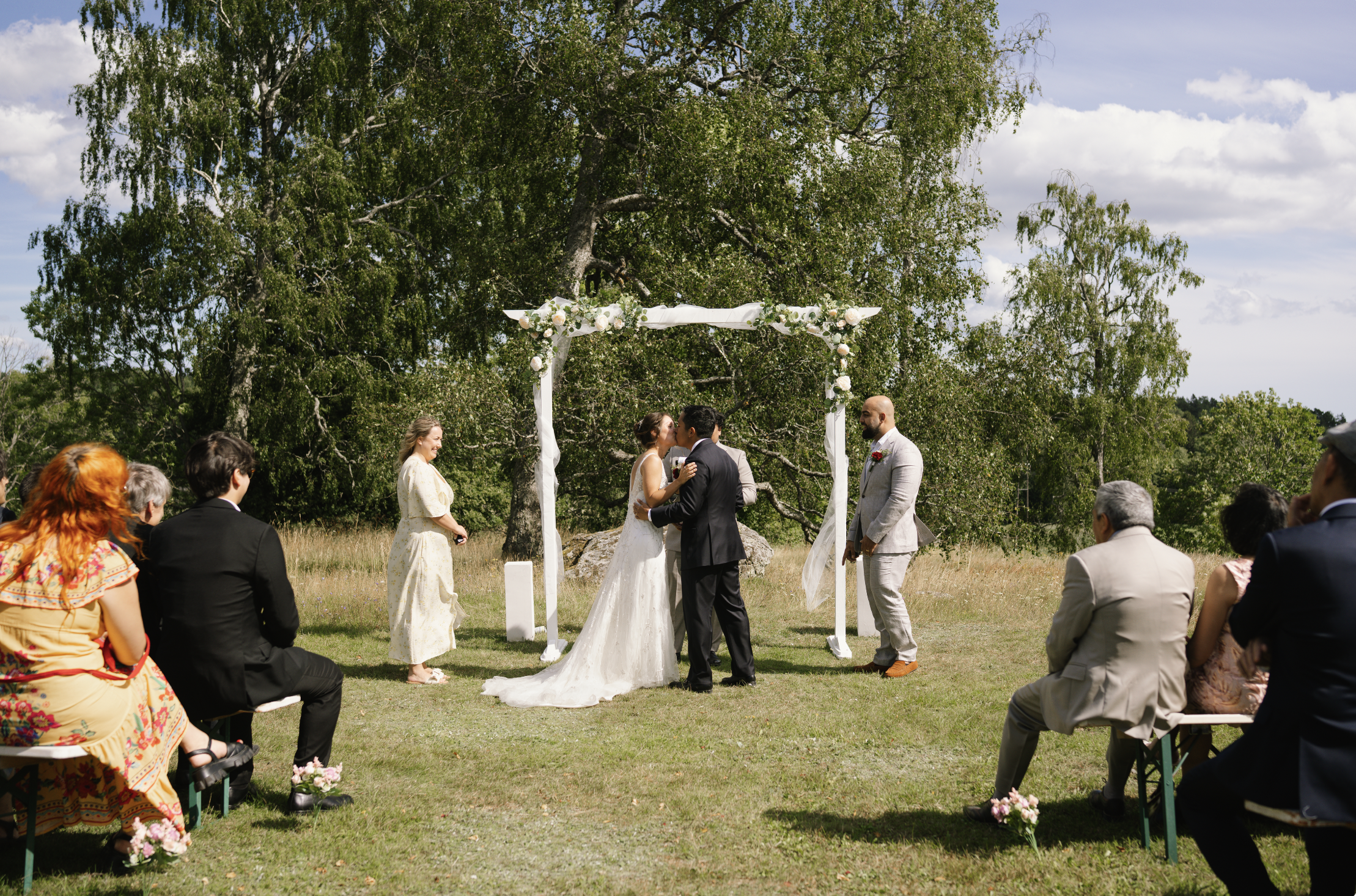 A couple sharing a kiss at an outdoor wedding ceremony under a white floral arch in a grassy field, with a large tree and blue sky in the background, surrounded by seated guests.