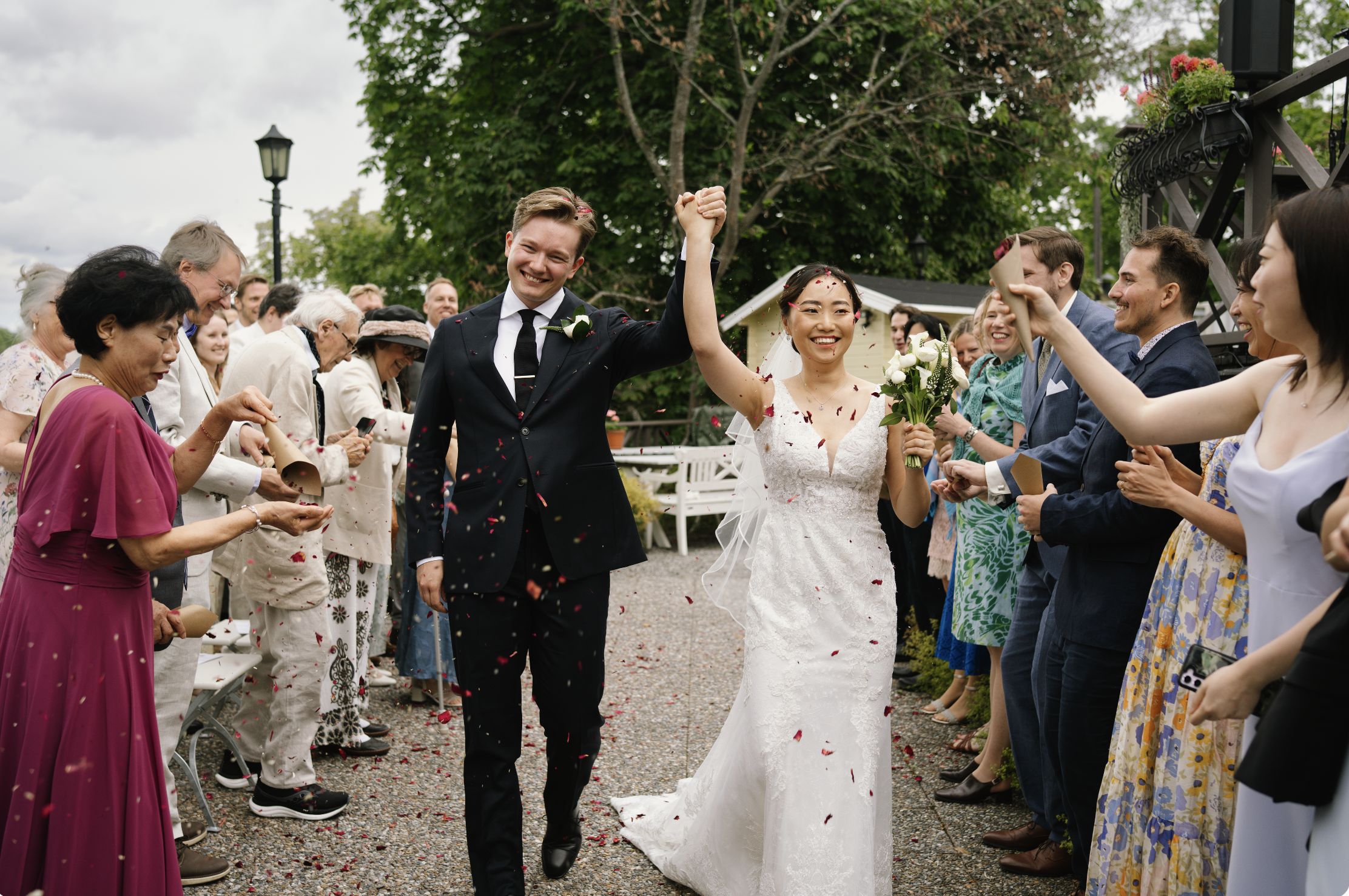 A newlywed couple walking arm-in-arm through a crowd in Stockholm at an outdoor wedding celebration, smiling and waving, with guests throwing confetti around them capture by swedish wedding photographer.