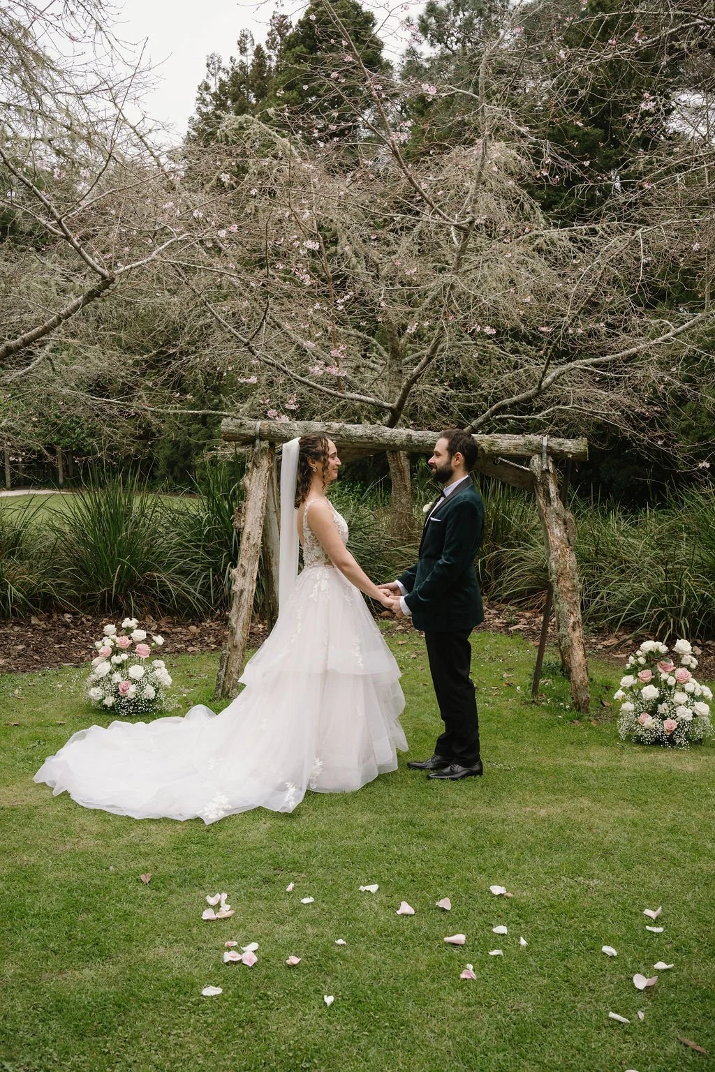 A bride and groom holding hands and looking at each other during their wedding ceremony outdoors, under a wooden arch with pink flowering trees in the background, with flower arrangements on either side and rose petals on the grass