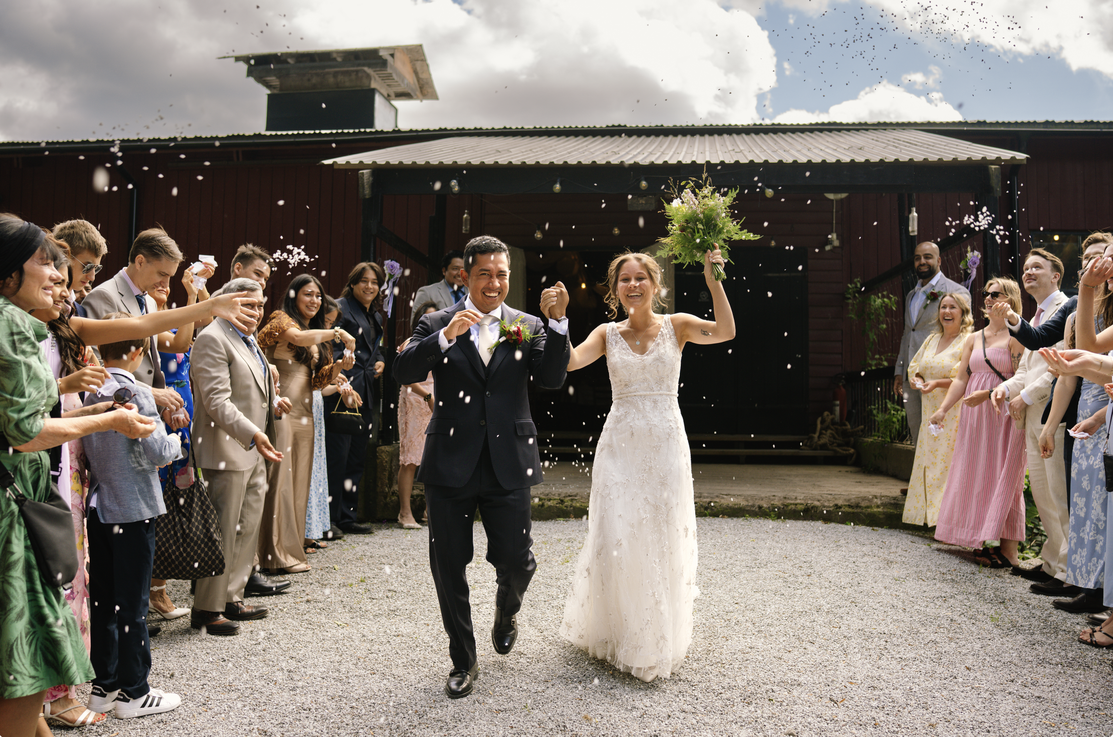 A newlywed couple walking joyfully through a crowd outside a rustic building, with guests celebrating and throwing confetti in the air.