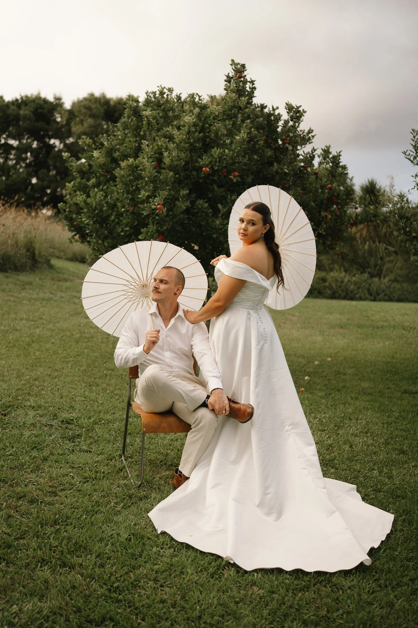 A man and woman in wedding attire with parasols outside near a tree.