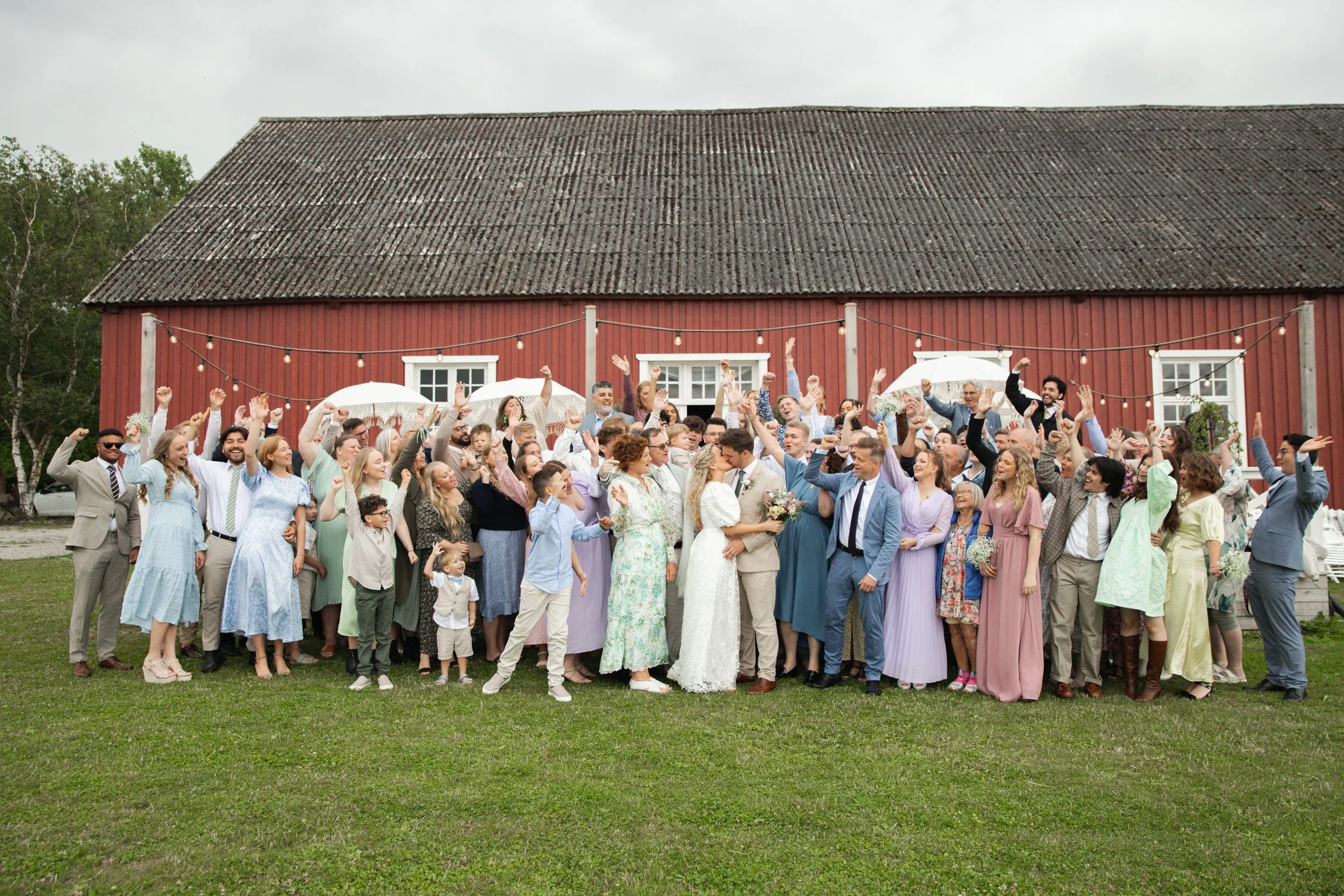 A large group of people at a wedding celebration, standing on a lawn in front of a red barn with string lights, some holding umbrellas.