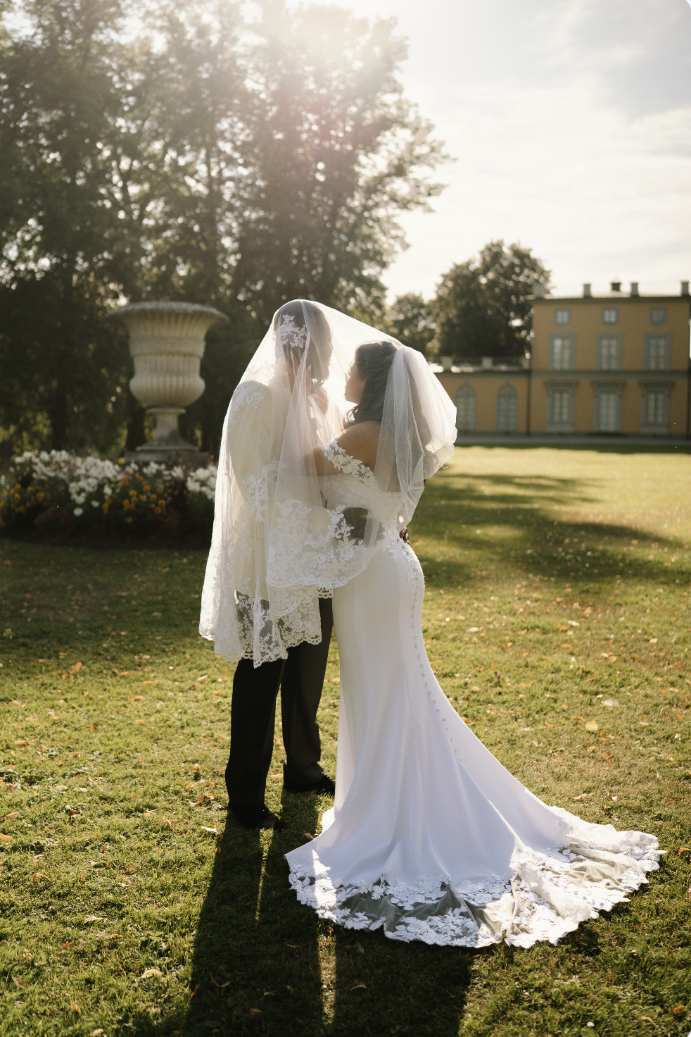 Two brides in wedding dresses under a veil, standing close and touching foreheads, outdoors on a sunny day with trees and a yellow building in the background.