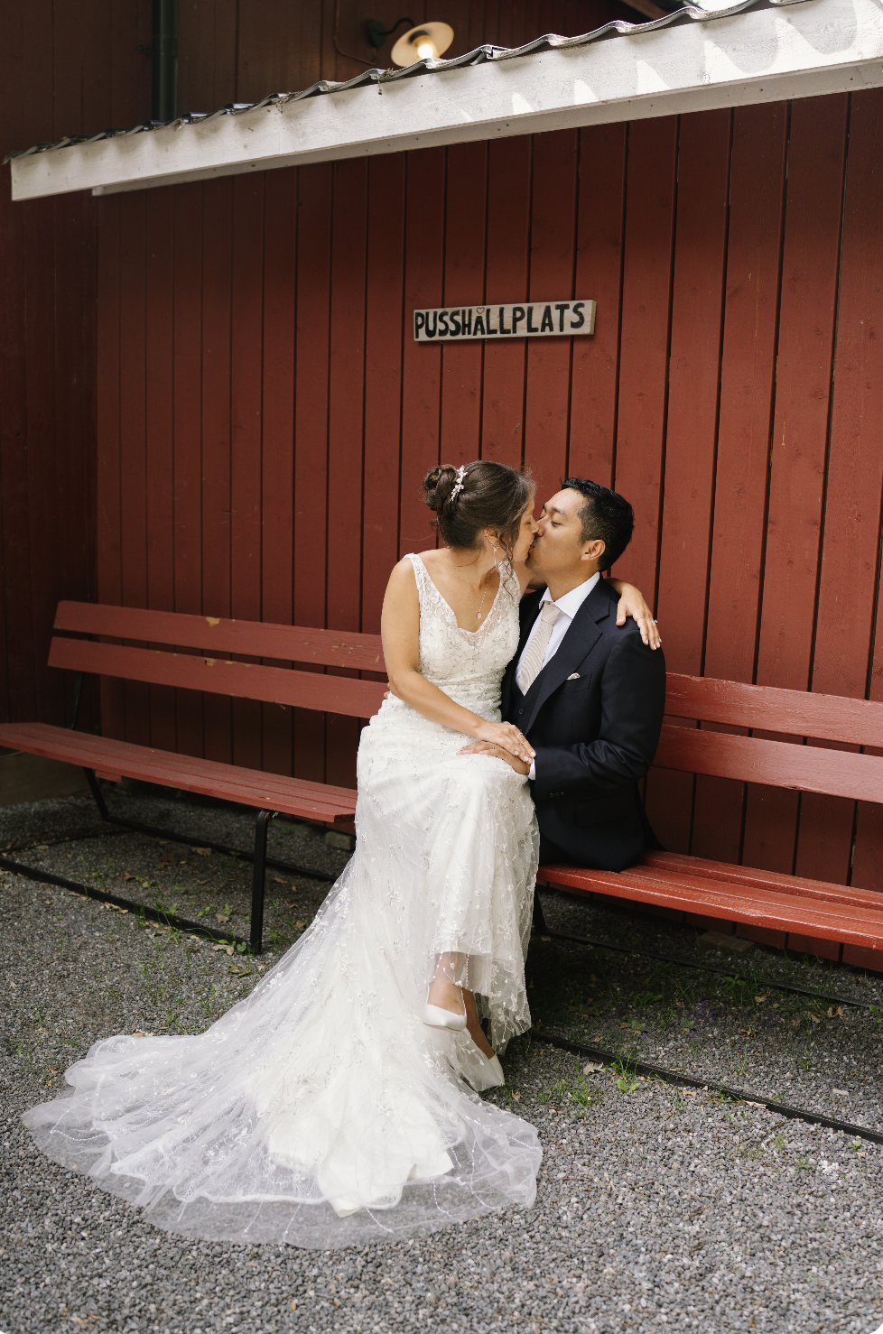 A bride and groom sharing a kiss in Stockholm on a red wooden bench against a red wooden wall. The bride is wearing a white lace wedding dress, and the groom is dressed in a dark suit captured by swedish wedding photographer