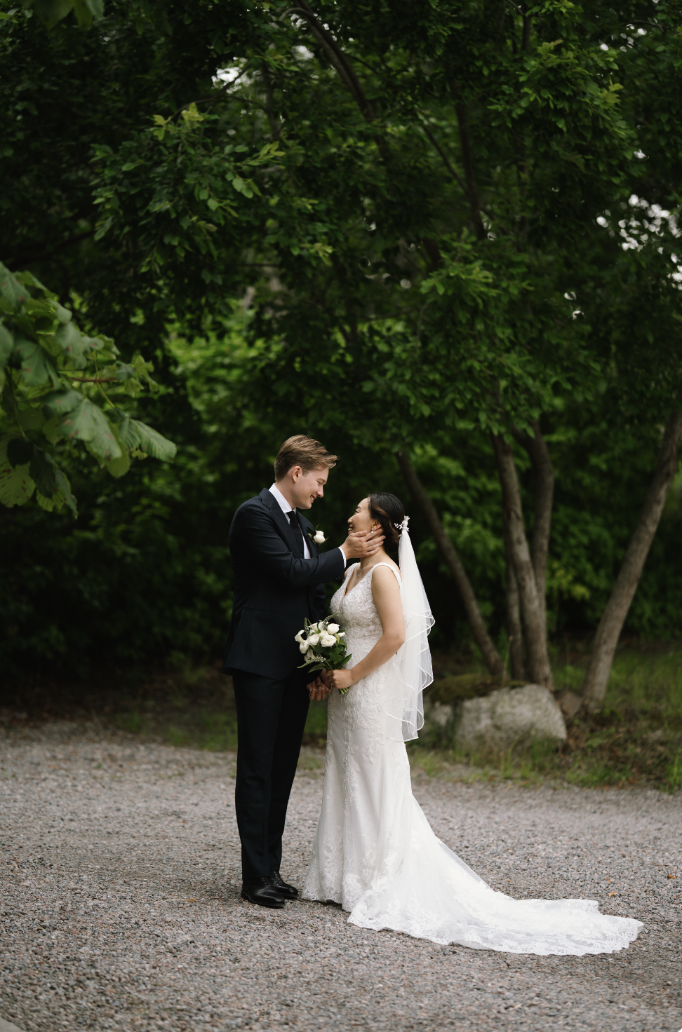 A newlywed couple standing outdoors, with a lush green background, the groom in a black suit holding the bride's face, and the bride holding a bouquet of white flowers, both smiling at each other.