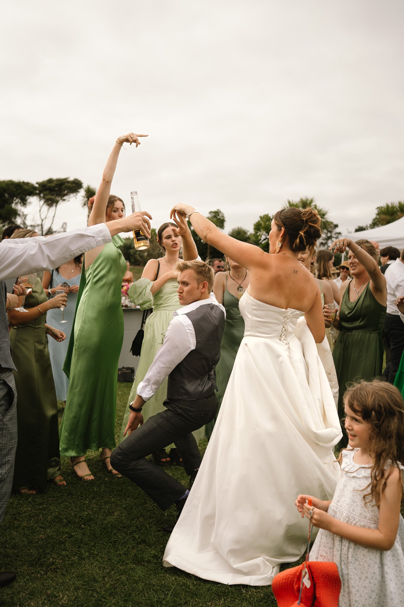 People dancing and celebrating outdoors at a wedding reception, with the bride in a white wedding gown and others in green and other colored dresses.