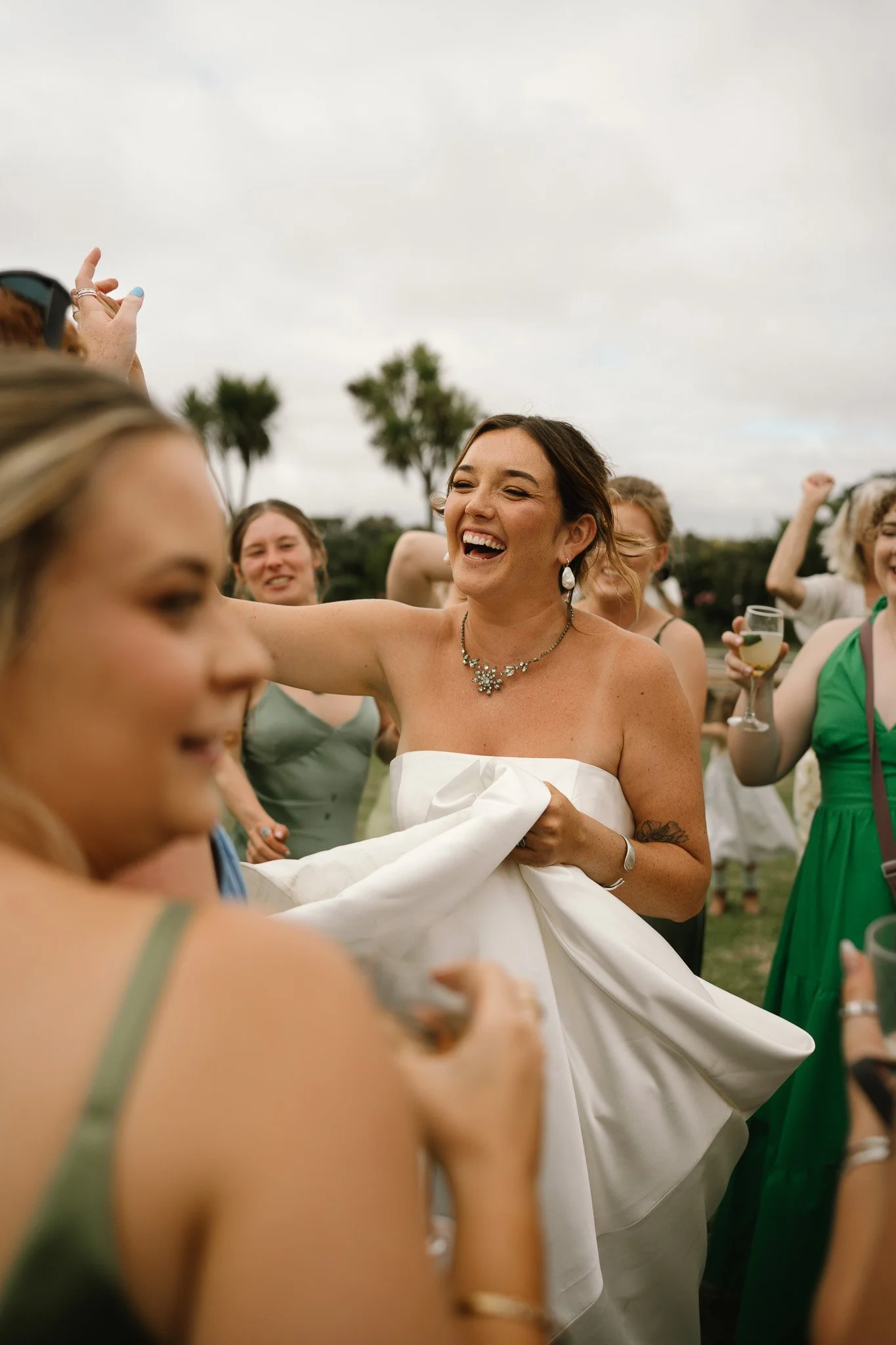 A bride in a strapless white wedding dress, smiling and holding her dress, surrounded by women celebrating outdoors, some holding drinks.