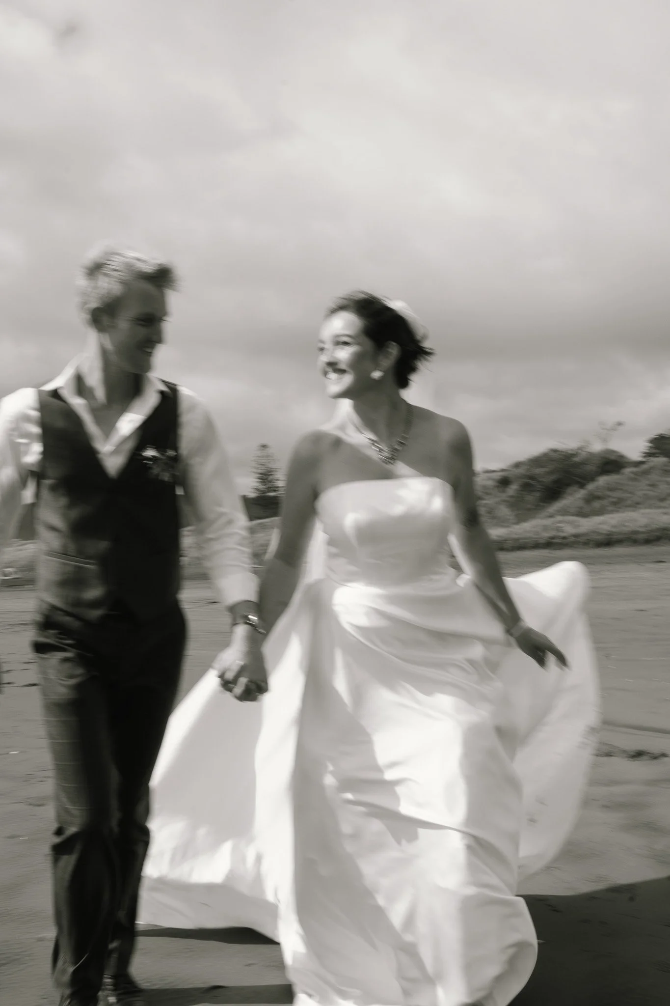 A black and white photo of two women holding hands, walking on a beach; the woman on the right is wearing a strapless wedding dress, and the woman on the left is dressed in a vest and shirt, suggesting they are a same-sex couple celebrating their wed
