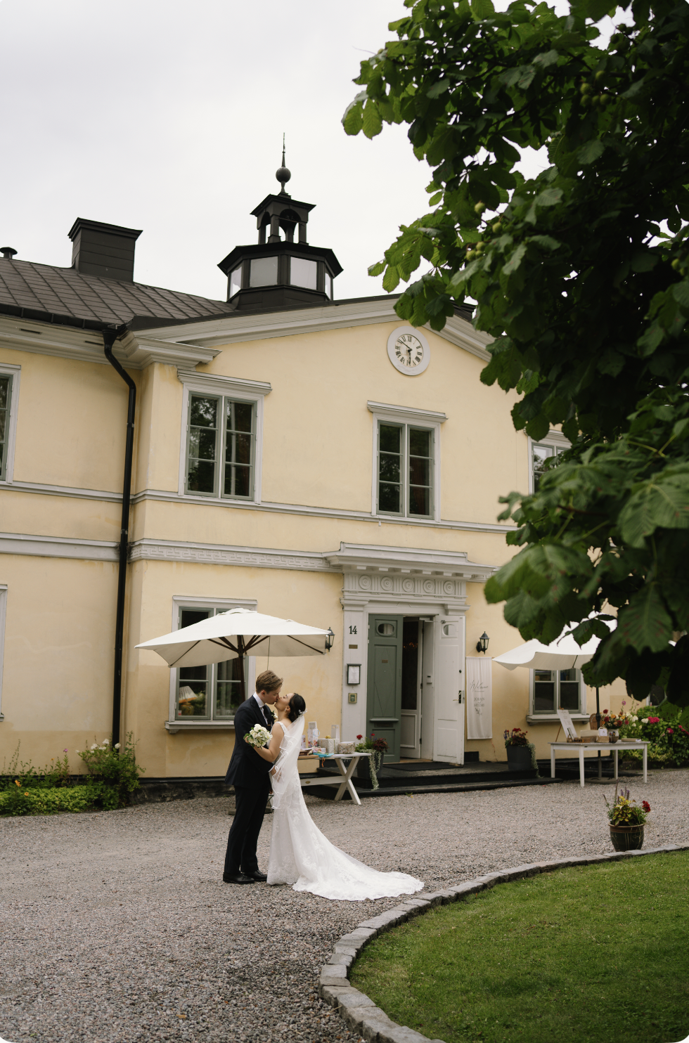 A bride and groom sharing a kiss outside a yellow slott in Stockholm with a clock on the wall, umbrellas, tables, and greenery.