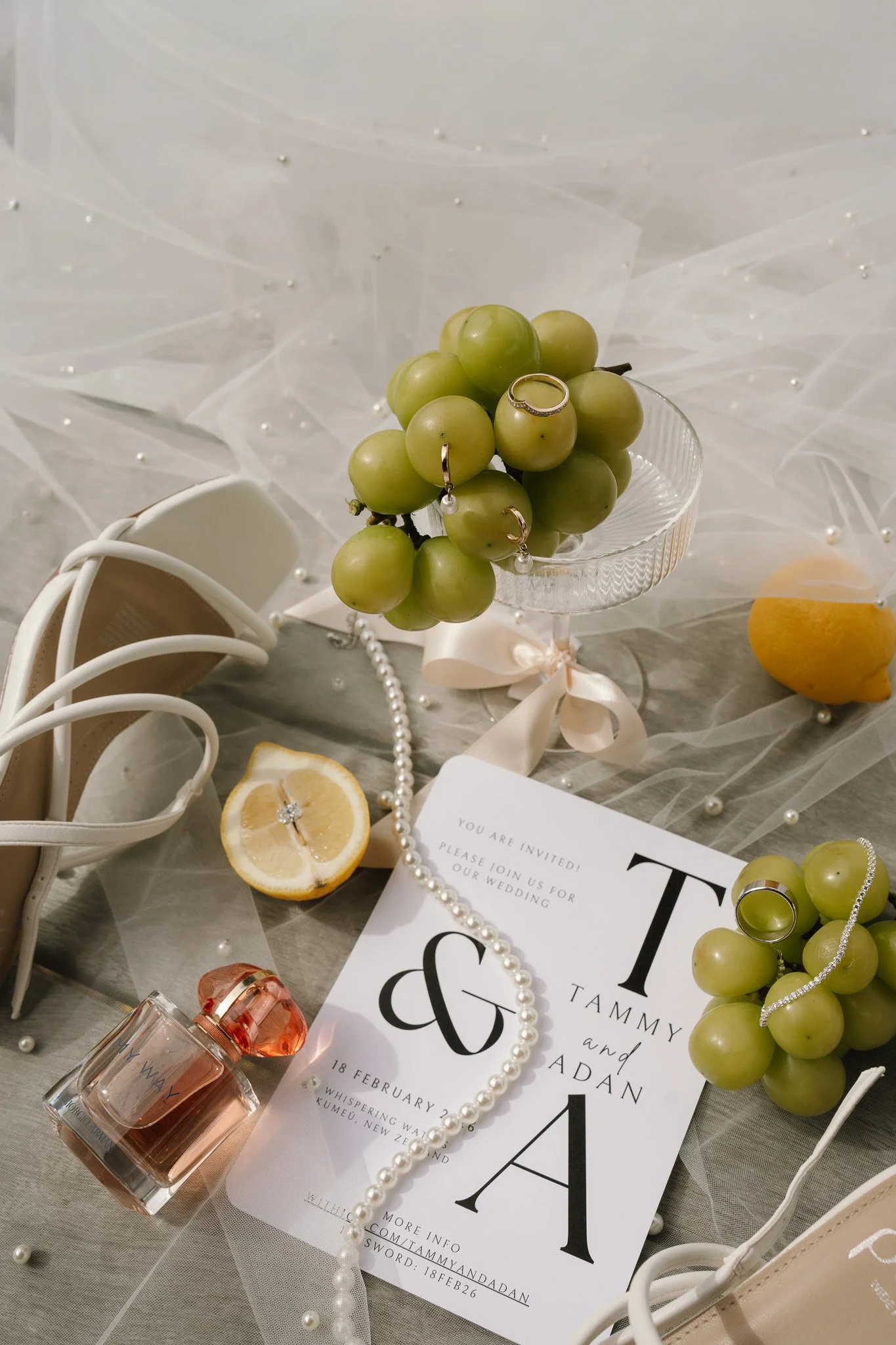 Wedding invitation surrounded by grapes, lemon slices, a pearl necklace, a perfume bottle, wedding rings, a pair of shoes, and decorative pearls on a sheer fabric background.