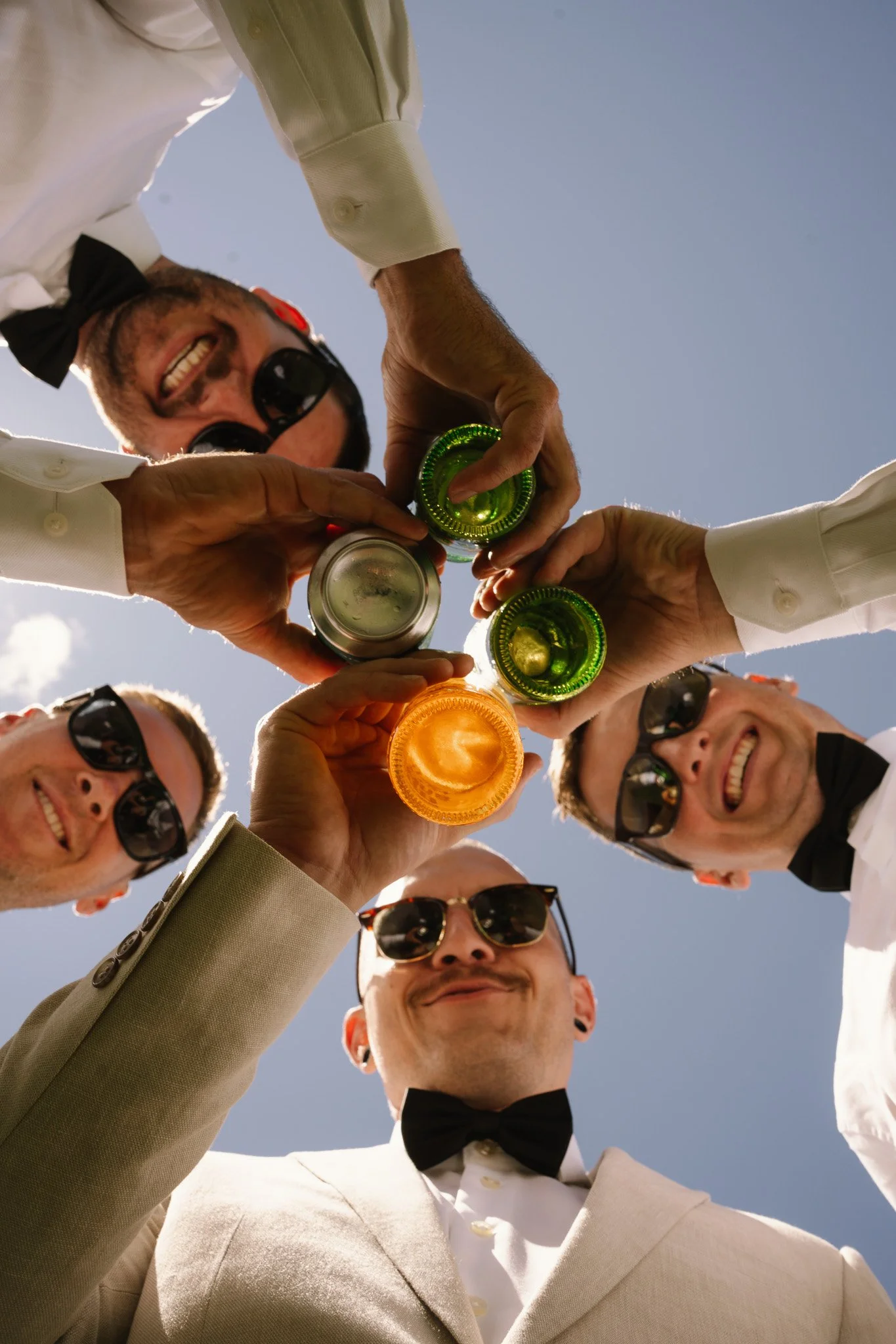 Group of men in formal attire wearing sunglasses, smiling, and raising colorful glasses in a toast under a clear blue sky.