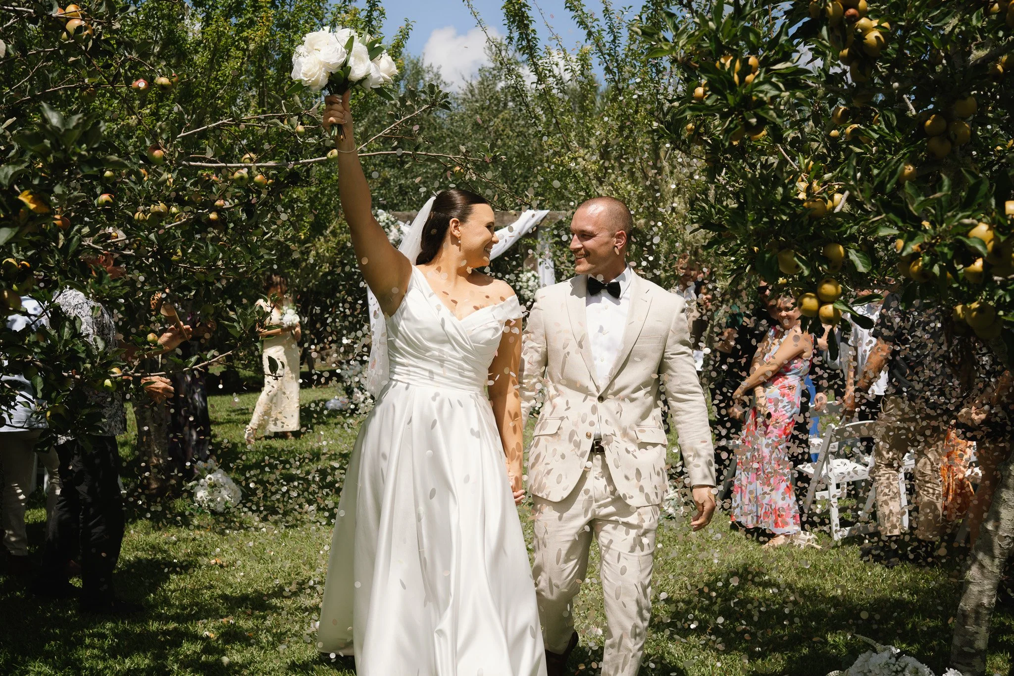A newly married couple walking hand in hand through a garden with apple trees, with guests throwing flower petals and celebrating.