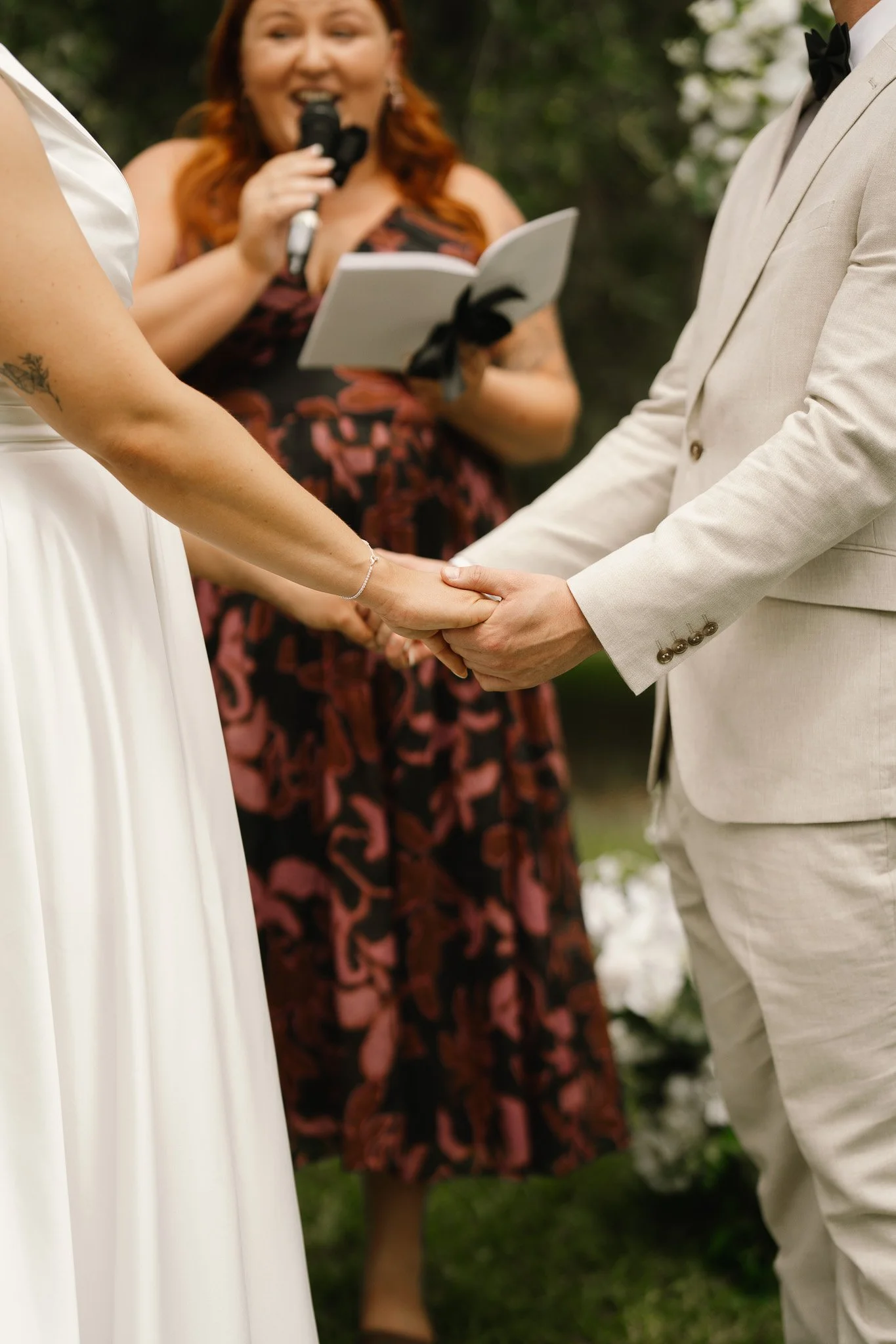 A couple holding hands during their wedding ceremony, with a woman reading vows or officiating, in an outdoor setting.