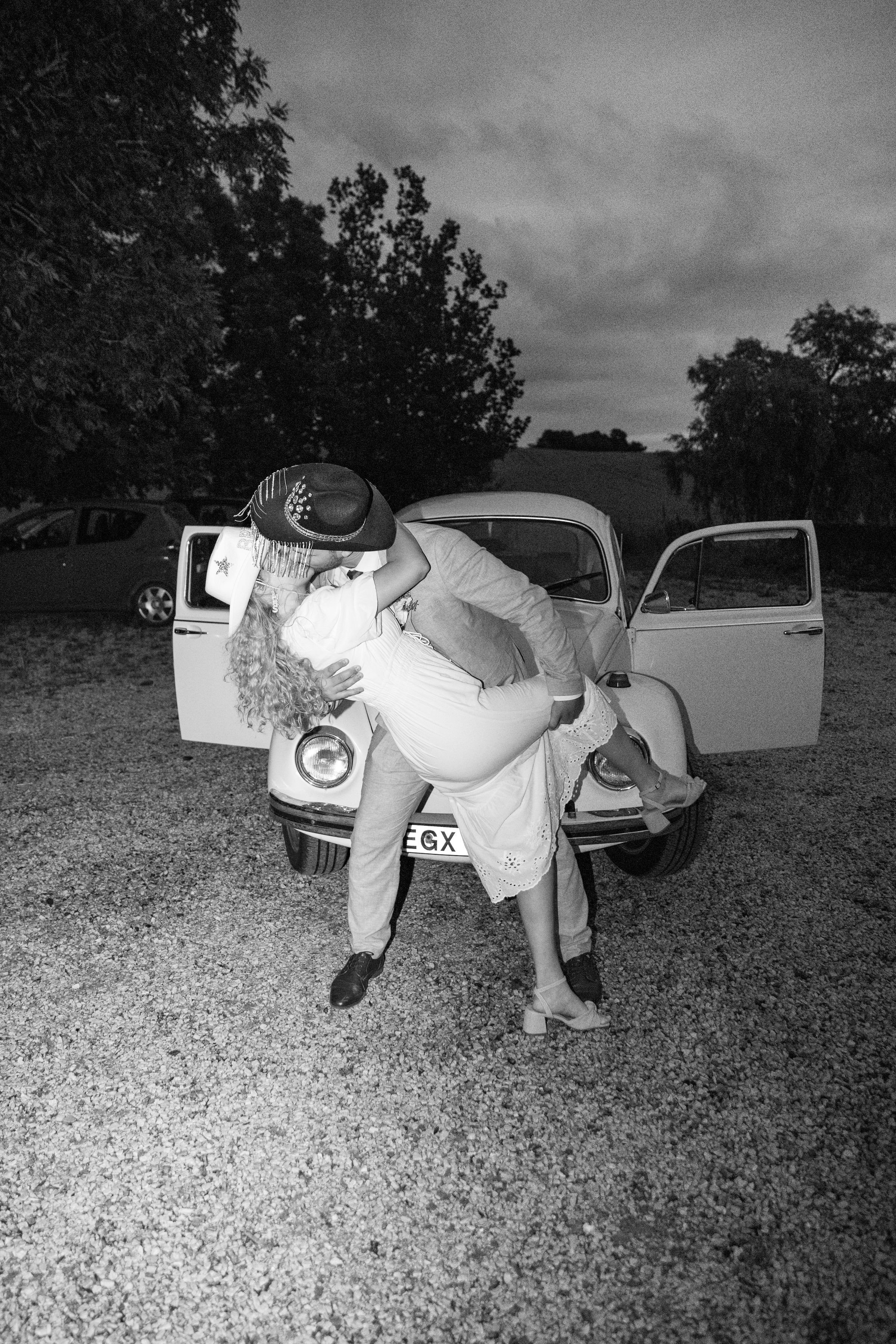 A black-and-white photo of a couple dancing in front of a vintage car, with trees and a cloudy sky in the background.