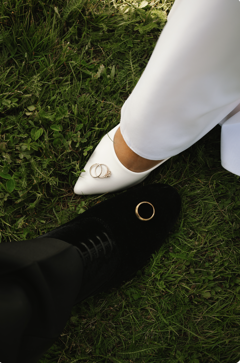Close-up of two people's feet on grass, one wearing a white pointed shoe with rings on top and the other in black ankle boots with a ring on the shoe.