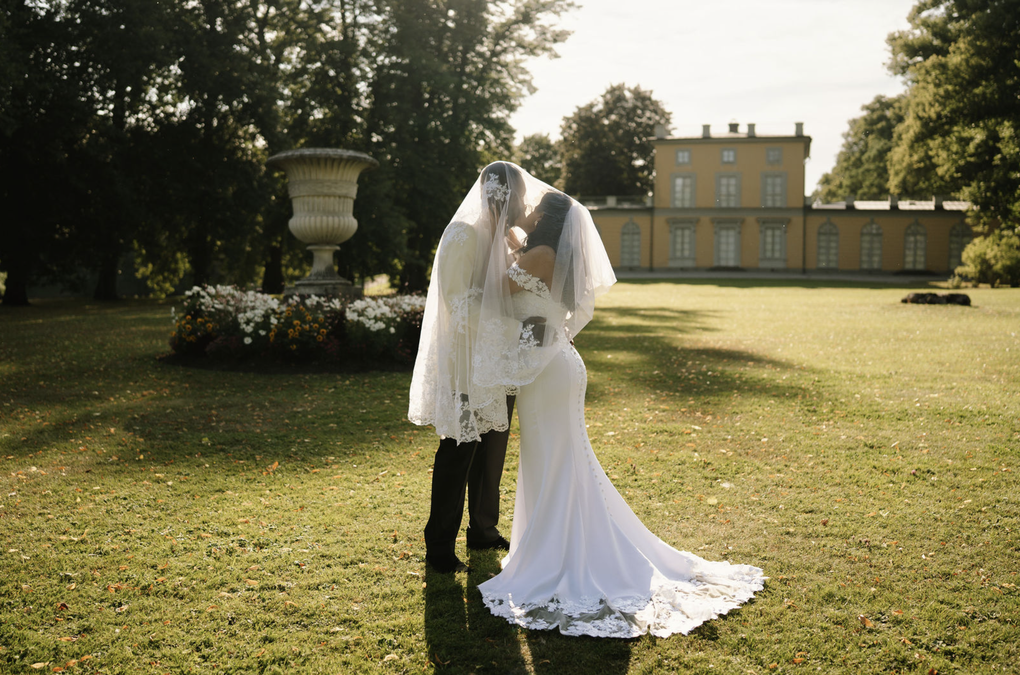 A bride and groom sharing a kiss outdoors in front of a historic yellow building and large urns, with the bride wearing a white dress and veil, and the groom in black tuxedo, during sunset.